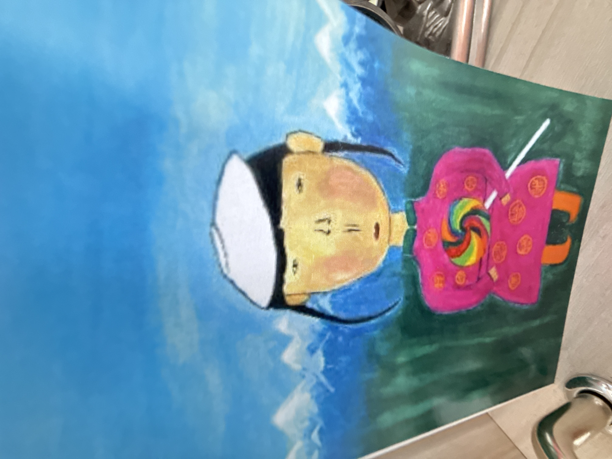 The image depicts a child's drawing of a character wearing a white hat and a pink robe, holding a rainbow lollipop. The background features a blue sky with white mountains and green land. The drawing is on a white surface, potentially paper or canvas, and appears to be photographed indoors. The location in the city of Unknown, Unknown is not applicable as this is a depiction and not a real-world scene.