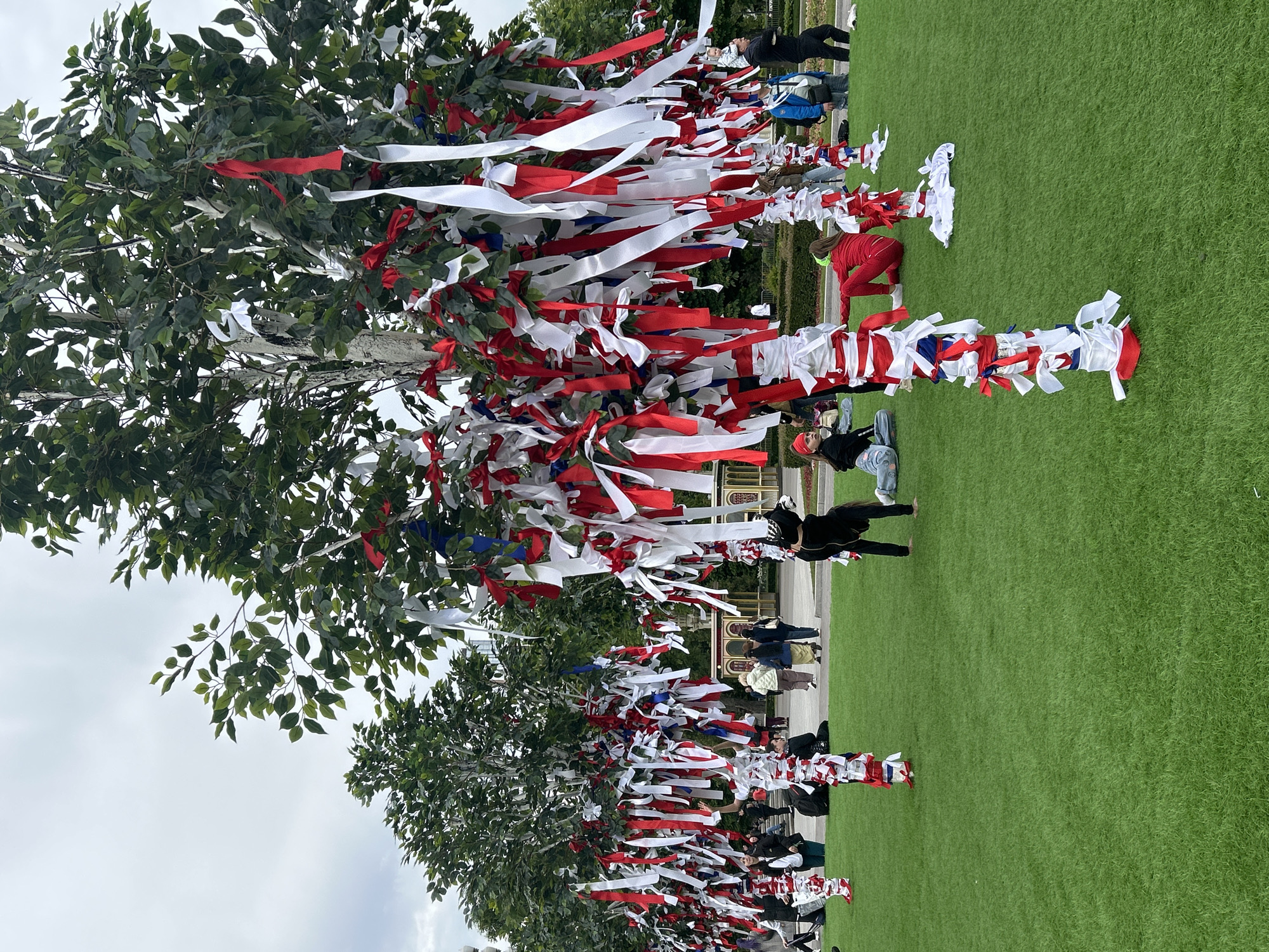 A sideways wide shot depicts a public gathering in what appears to be a park, possibly in Moscow, Russia. A row of trees is adorned with red, white, and blue ribbons, resembling a makeshift memorial or decorative display. Several people are standing and sitting on the green lawn, some interacting with the ribboned trees. The background shows glimpses of buildings and architecture, contributing to the impression of an urban park setting. The sky is overcast, suggesting daylight conditions.