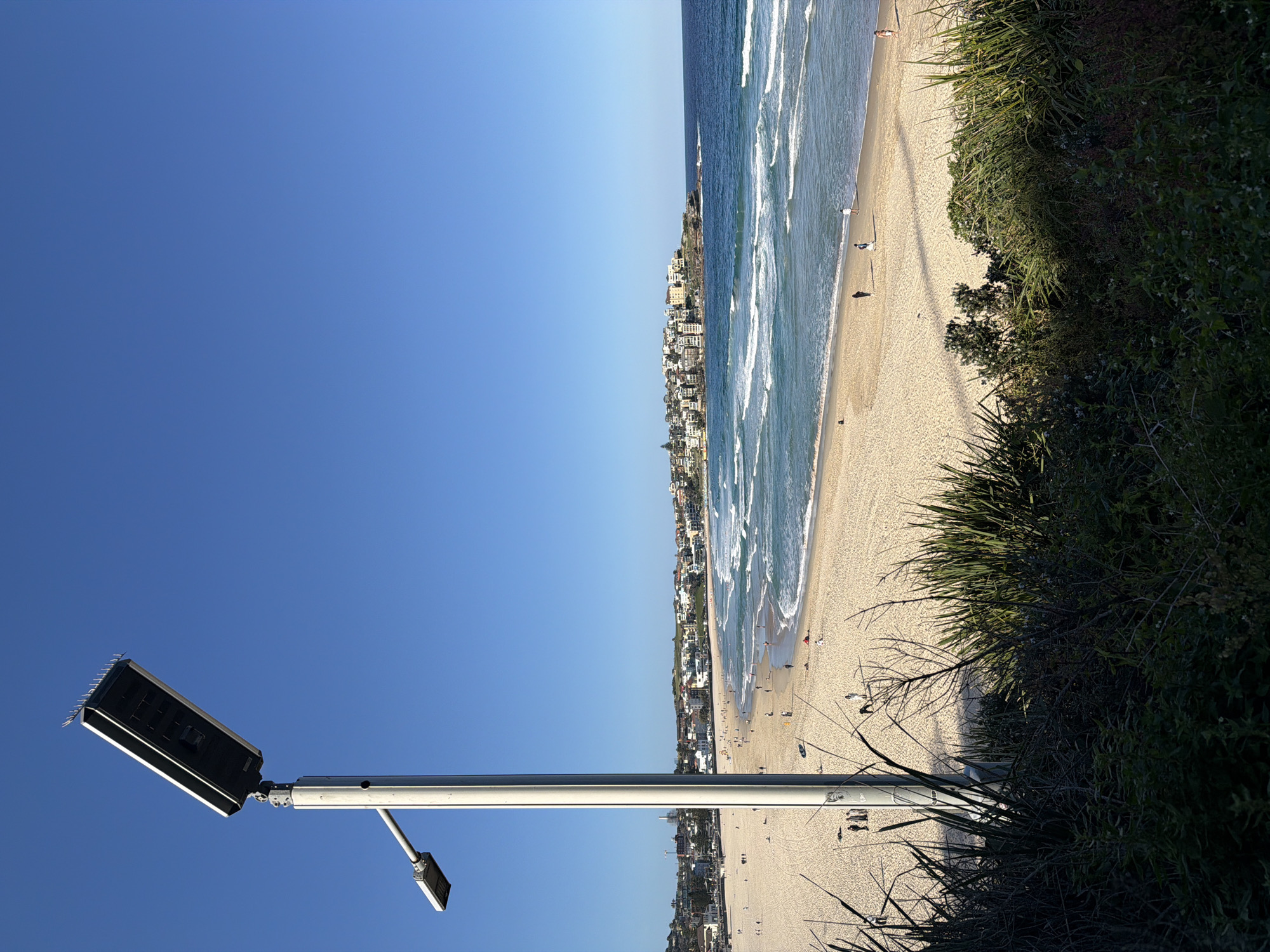 The image captures a sunny day at Bondi Beach, Australia, with a clear blue sky overhead. A modern solar-powered street lamp stands prominently in the foreground, its long pole extending upwards. To the left of the main lamp, a smaller, secondary light fixture is visible on an extended arm. The beach itself is wide and sandy, with gentle waves rolling onto the shore. Several individuals are scattered across the sand and in the water, some walking, others appearing to relax. In the background, a cluster of buildings lines the coastline, characteristic of a populated beachfront area. Lush green vegetation occupies the bottom of the frame, partially obscuring the view of the beach below.