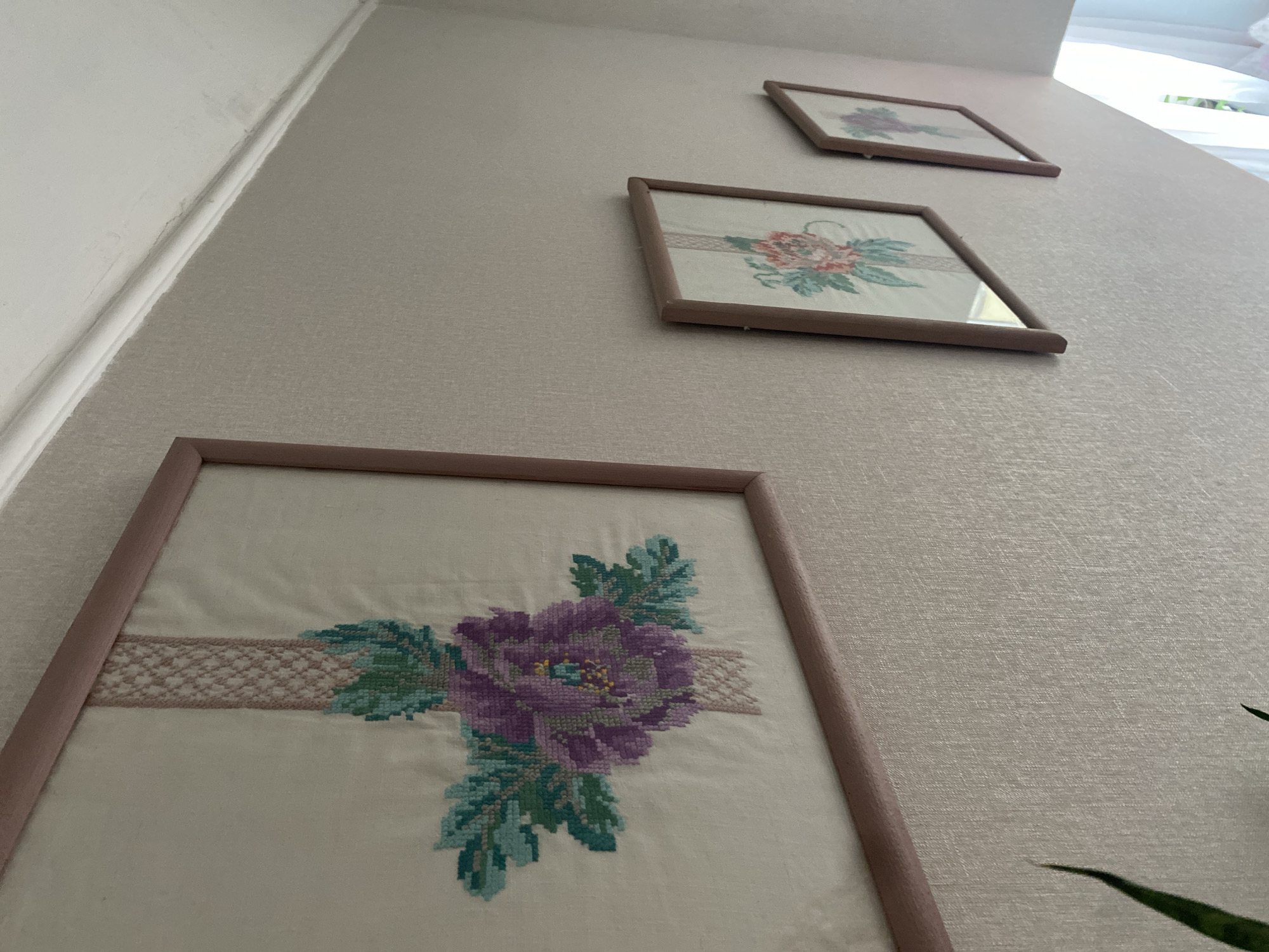 The image presents an upward-angled view of an interior wall adorned with three framed cross-stitch artworks. The wall is covered in a light beige or greige wallpaper with a subtle vertical texture. A white ceiling with crown molding runs along the top left edge of the frame.
The three rectangular frames, each with a light brown or pale pink wooden border, are arranged diagonally upwards along the wall. The closest frame, prominent in the foreground, displays a detailed cross-stitch of a vibrant purple flower, possibly a peony or rose, with accompanying green and teal leaves. A light pink, intricately patterned horizontal band is stitched across the fabric behind the main floral motif.
Further up the wall, a second framed picture showcases a distinct cross-stitch design featuring pink and green floral elements. The third and smallest frame is positioned highest and to the right, exhibiting a less clear but consistent floral cross-stitch pattern.
Soft, natural light, originating from an unseen window or light source in the upper right, illuminates the scene, suggesting it is daytime. A small portion of a green plant leaf is visible in the bottom right corner of the original image, implying other decorative elements nearby. No specific activity is taking place, and no text is visible in the image.