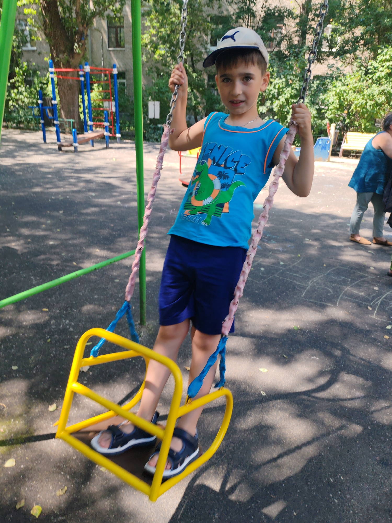 The image depicts a young boy on a yellow swing set at a playground. He is wearing a blue tank top with a dinosaur graphic and blue shorts, along with a white and blue baseball cap. The swing set has green poles and chains with pink and white rope. In the background, there is a red and blue climbing structure and trees. To the right, a person in a blue top and grey pants is visible, and another person's legs and feet are in the lower right corner. The ground is asphalt with chalk drawings, suggesting it is a public park or schoolyard. The presence of a multi-level climbing structure with blue and red bars, typical of Soviet-era playground equipment, suggests the location is likely in Moscow, Russia, possibly in a residential courtyard or a small public park. The overall atmosphere is casual and sunny.