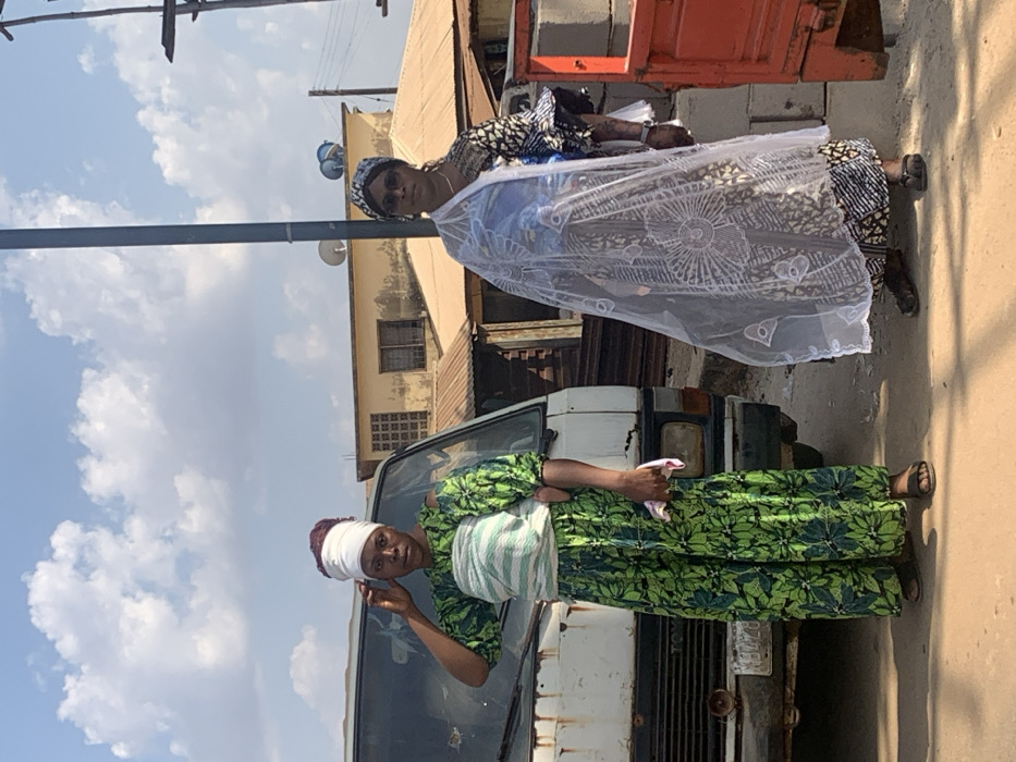 Two women stand on a street next to a dilapidated white van. The first woman wears a green floral dress with a white head wrap and sash. She is positioned in front of the van, holding a pink item. The second woman, located further back, wears a patterned blue dress, a matching head covering, and sunglasses. A white lace shawl drapes over her shoulders. The background features buildings under a partly cloudy sky. A utility pole stands to the left.