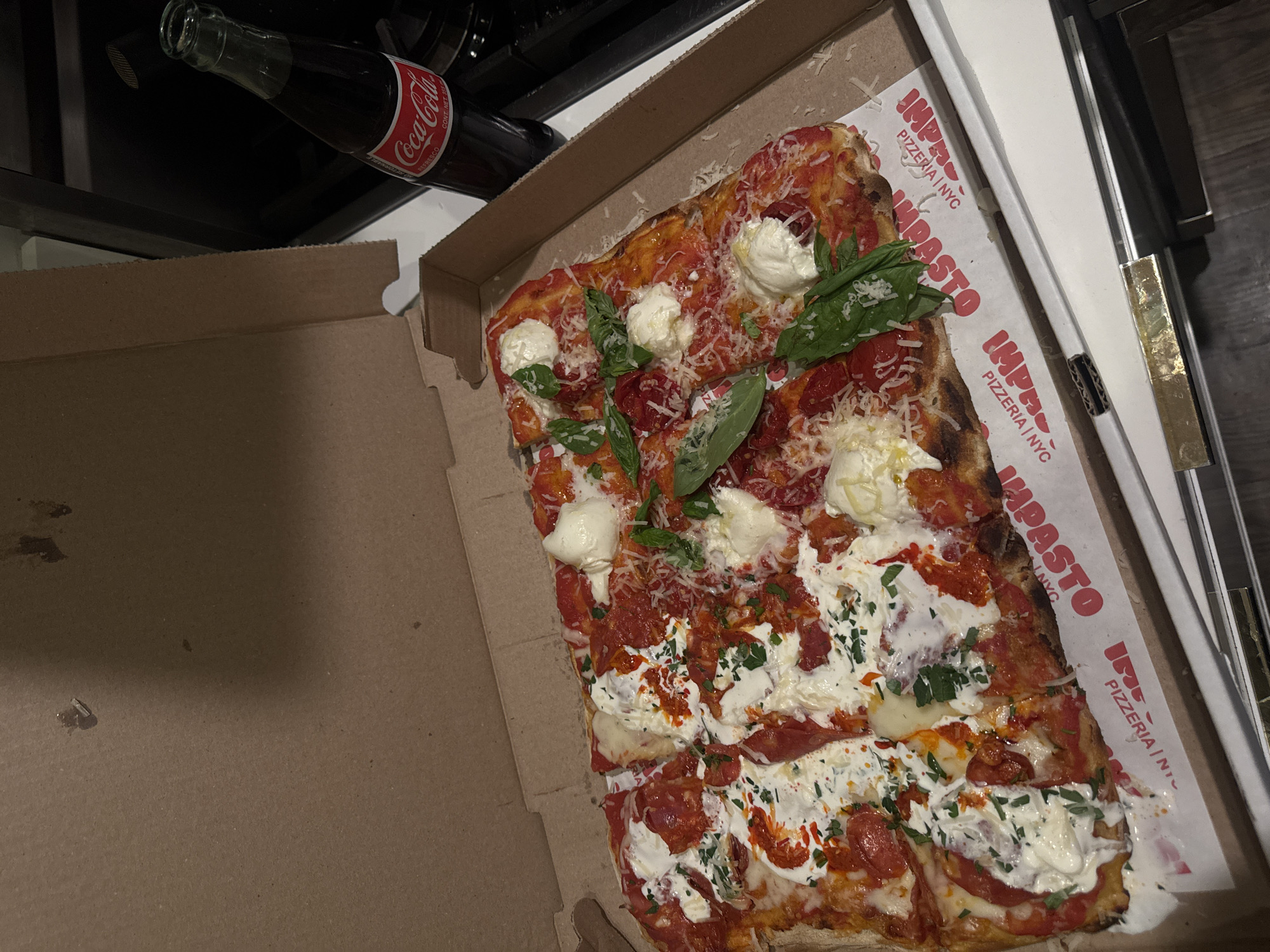 This is an overhead shot of a rectangular pizza served in a brown cardboard pizza box, resting on a light-colored countertop. The pizza features a tomato sauce base, melted white cheese, multiple dollops of fresh white cheese (likely burrata), slices of pepperoni, whole basil leaves, chopped green herbs (possibly parsley or oregano), a dusting of grated hard cheese, and a drizzle of red chili oil. The pizza is cut into multiple portions. The white paper lining inside the pizza box clearly displays "IMPASTO PIZZERIA | NYC" in red lettering, indicating the location in New York City, United States of America. To the right of the pizza box stands an uncapped, dark glass bottle of Coca-Cola, with its red and white label visible. In the background, a dark kitchen appliance, possibly a stove or oven, is partially visible. The scene is illuminated by indoor lighting.