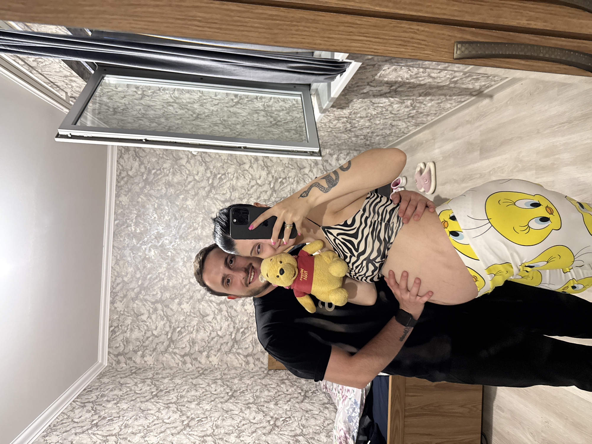 A pregnant woman and a man are posing for a mirror selfie in what appears to be a bedroom. The woman is wearing a black and white zebra-print top and yellow pants with a Tweety Bird pattern. She is holding a Winnie the Pooh plush toy. The man is wearing a black t-shirt and is standing behind the woman, with his arms around her. The room has textured wallpaper, a wooden floor, and a large mirror that reflects the scene. A window with grey curtains is visible in the background. A wooden dresser or bed frame is also present. There are pink sandals on the floor. The image captures a moment of anticipation and joy, likely in a domestic setting.