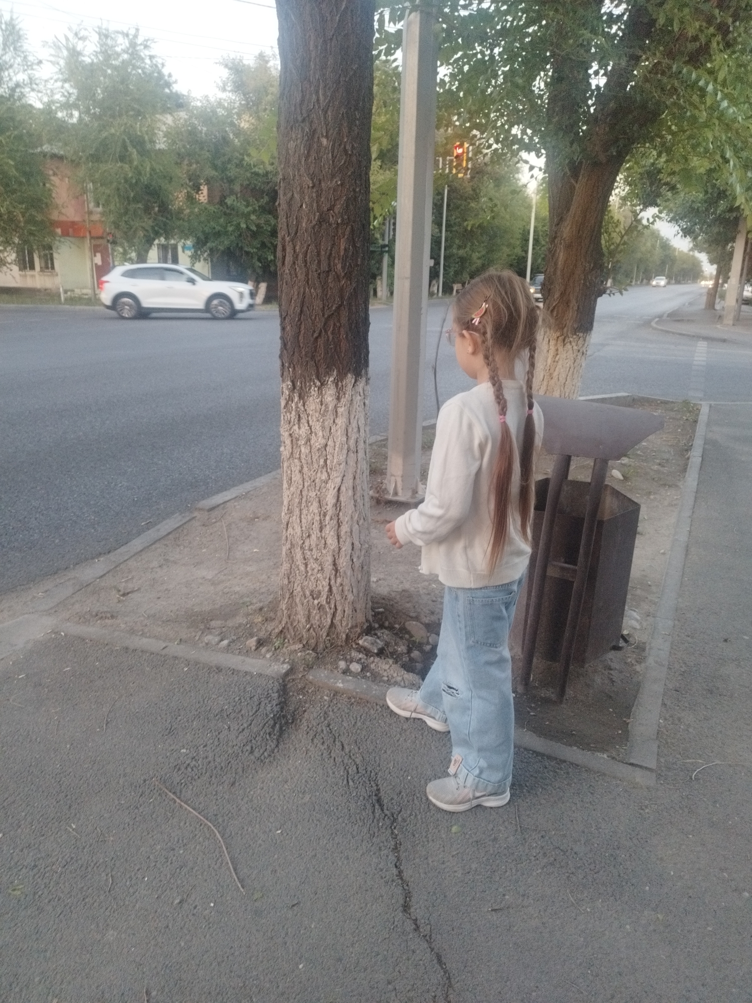 The image depicts a young girl with blonde hair in two braids, wearing a light-colored sweater and ripped denim jeans, standing on a sidewalk next to a tree. She is facing away from the camera, looking towards a street. In the background, a white SUV is visible driving on the road, and beyond that, a building and trees line the street. A dark metal trash can is located near the girl. The scene appears to be in an urban environment, consistent with Shymkent, Kazakhstan. The lighting suggests it is daytime, possibly late afternoon or early evening.