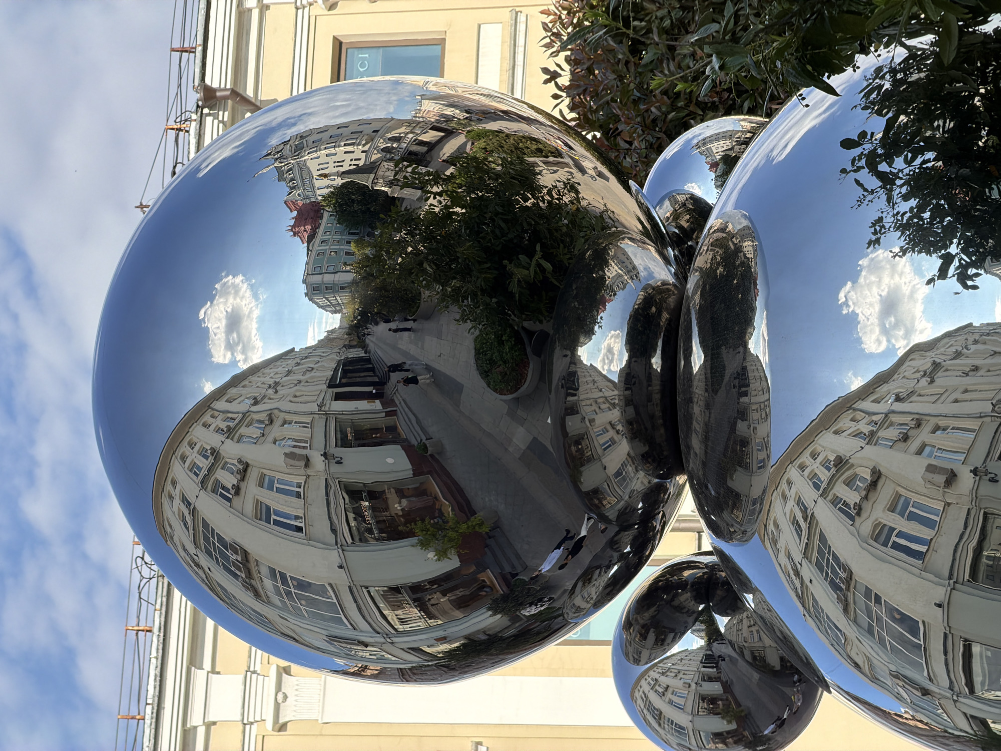 The image shows a cluster of large, highly polished, spherical sculptures reflecting the surrounding cityscape. The reflections include buildings with numerous windows, a street with people walking, trees, and a blue sky with white clouds. The sculptures are positioned outdoors, near a building with a beige facade. The reflections suggest that the location is Moscow, Russia, specifically near TSUM department store on Petrovka street, indicated by the reflection of the red building in the distance. The scene appears to be a sunny day. The sculptures are the primary subjects of the photograph.