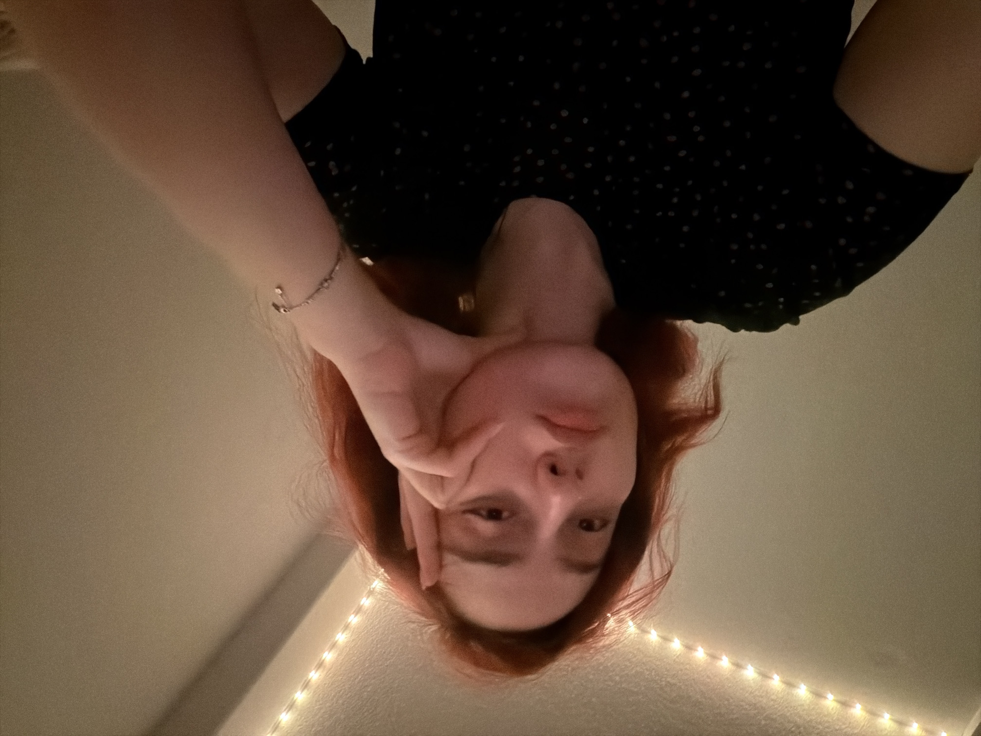 The image is a close-up, indoor, waist-up shot of a young woman with reddish hair. She is leaning her head on her hand, with her fingers splayed and resting on her cheek. Her expression appears pensive or bored, with her lips slightly pursed. She is wearing a black t-shirt with a subtle pattern of small dots. A string of small, warm-toned LED lights is visible along the top edge of the frame, suggesting she is in a room. A delicate silver bracelet with a small charm is worn on her left wrist. The background is a plain, light-colored wall. There is no discernible indication of the city of Strasbourg, France, or any other specific location within the image.