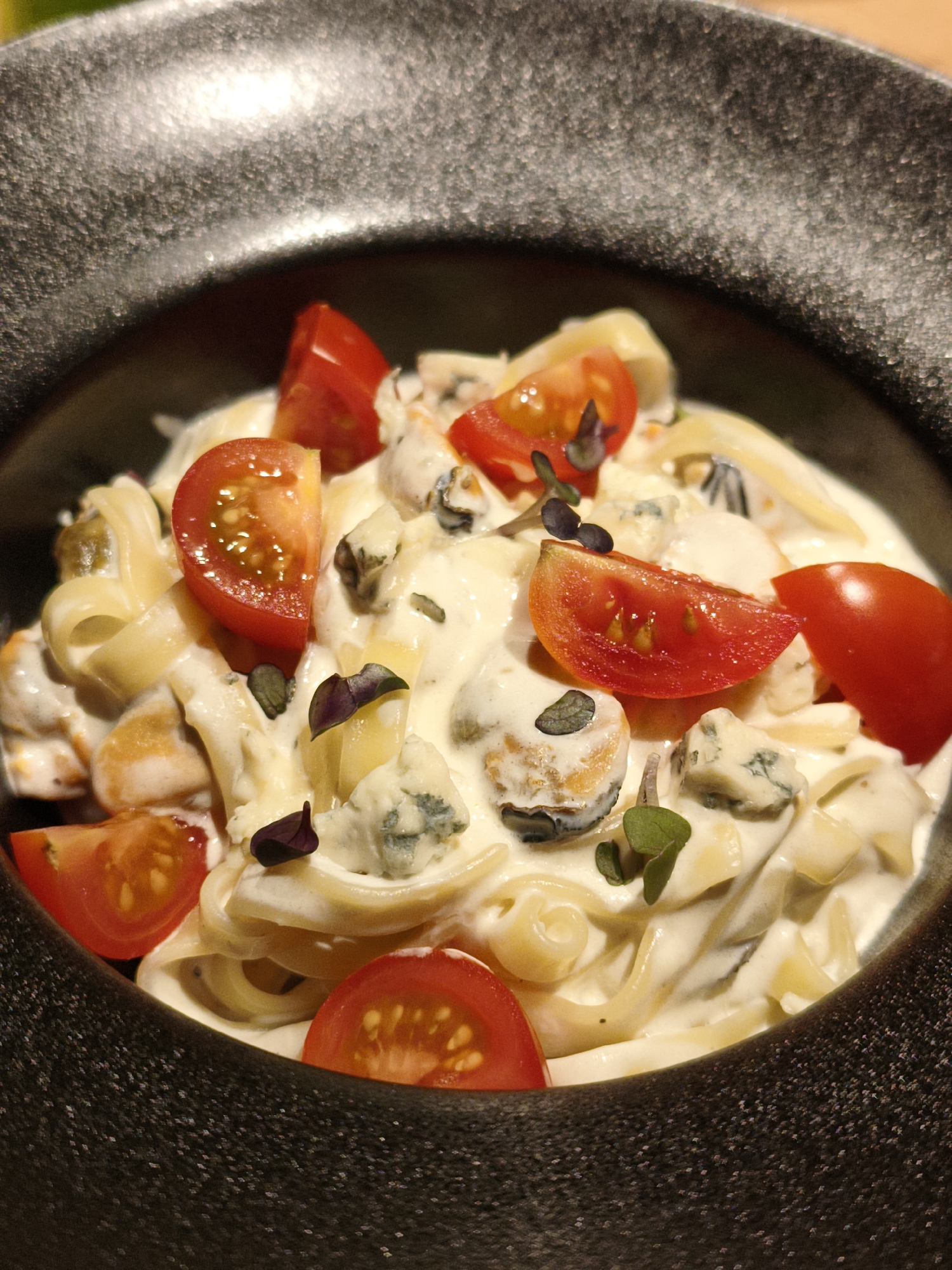This is a close-up, overhead shot of a pasta dish served in a dark, textured bowl. The pasta appears to be fettuccine or a similar flat noodle, coated in a creamy white sauce. Scattered throughout the pasta are pieces of blue cheese, halved cherry tomatoes, and what seem to be mussels. Small green garnishes, possibly microgreens, add a touch of color. The lighting suggests an indoor setting, likely a restaurant, possibly for a meal. There are no people visible in the frame, and no text is present. The overall impression is one of a rich and savory meal. The setting is Chaikovsky, Russia.