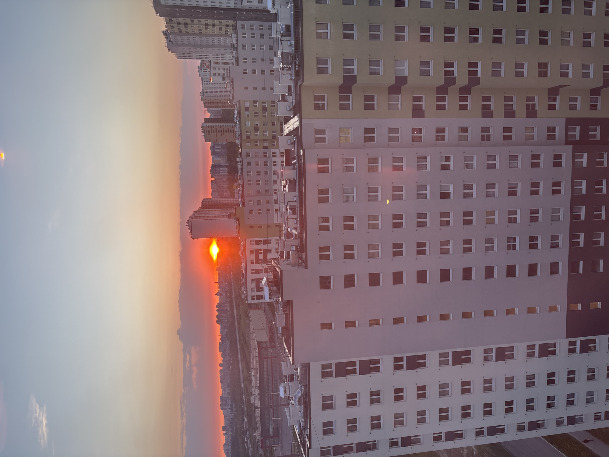 The image depicts a sunset over an urban landscape in Vsevolozhsk, Russia. Multiple high-rise buildings with repetitive window patterns dominate the foreground, with different colors like light yellow, white, and dark brown. In the distance, the setting sun is partially obscured by the city's horizon, creating a reddish-orange glow in the sky. Below the sun and buildings, the landscape features a mix of structures and vegetation. The sky transitions from a deep red near the horizon to a lighter orange and eventually pale blue higher up. A bright orb, potentially lens flare, is visible in the top left corner.