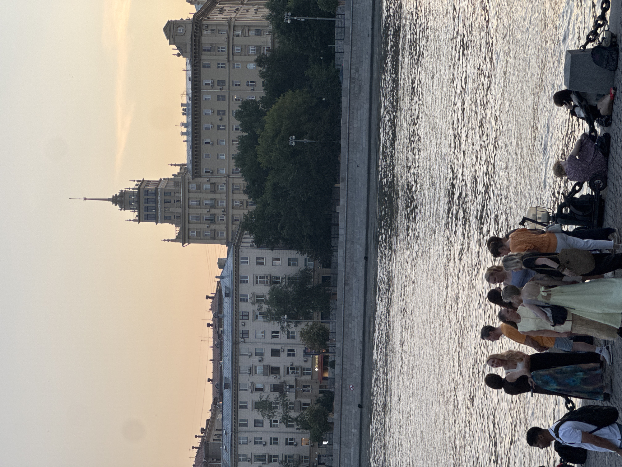 This media file depicts a waterfront scene in Moscow, Russia, at sunset. The perspective is vertical, suggesting the image was taken with the camera rotated.
In the foreground, a group of people are gathered along the embankment of a river, with their backs mostly to the viewer. They are dressed in casual summer clothing, and their postures suggest they are looking out at the water or a distant view. One person in a light orange shirt appears to be taking a photo with their phone. Another individual in a light green dress stands with their arms crossed. The overall mood seems relaxed and contemplative.
The river itself occupies a significant portion of the right side of the frame, its surface rippling and reflecting the soft, warm light of the setting sun. The embankment is constructed of stone blocks, and a heavy chain is visible, likely part of the mooring system.
In the background, a row of imposing, Stalin-era buildings lines the opposite bank of the river. These structures are characterized by their detailed architecture, numerous windows, and ornate tops. Trees are interspersed between the buildings, adding a touch of greenery to the urban landscape. The sky is a soft, hazy orange and yellow, indicative of twilight. The light suggests it is late afternoon or early evening. There are no overt signs of a specific event, rather it appears to be a typical scene of people enjoying a pleasant evening by the water in a city. No visible text is discernable in the image.