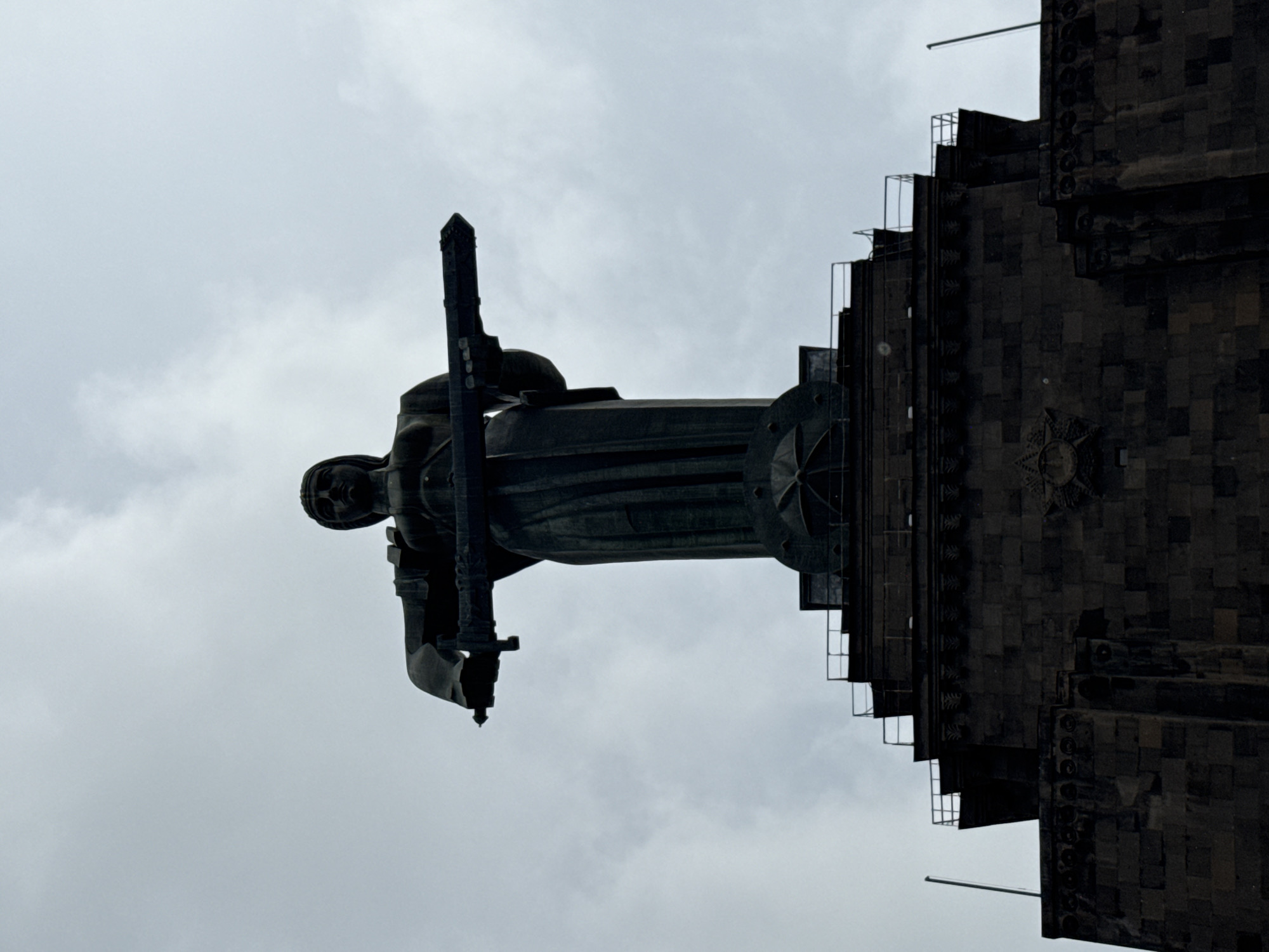 Looking up at the Mother Armenia statue, in Yerevan, Armenia, the statue stands on a tall pedestal attached to the Armenian Military Museum of the Ministry of Defense. The statue is of a female figure holding a sword. The sky is overcast. The photo is oriented sideways.