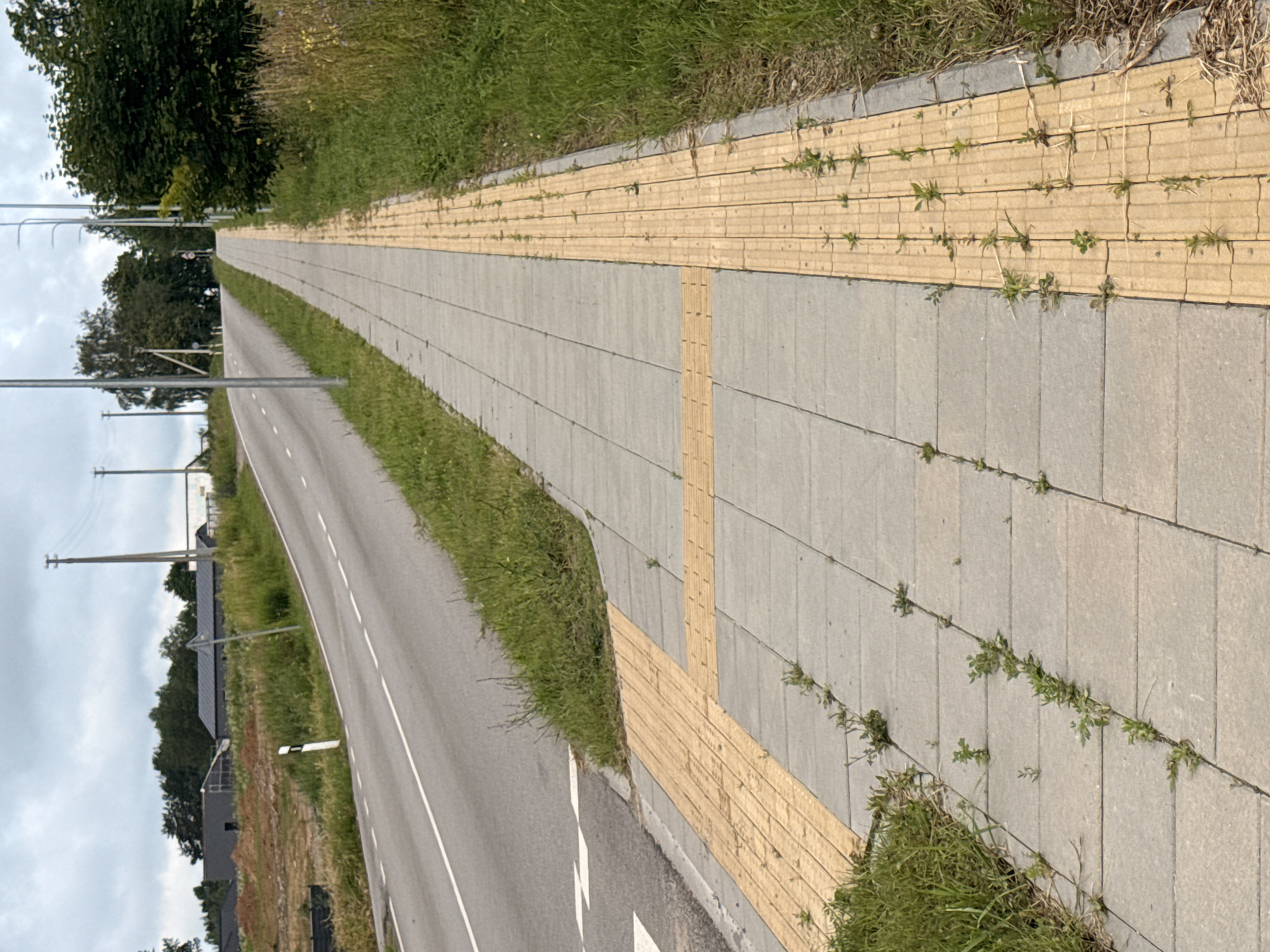 The image depicts a sidewalk alongside a road in Aukštieji Rusokai, Lithuania. The sidewalk is paved with light gray square tiles and features a tactile paving strip in a yellow color, designed for visually impaired pedestrians. The yellow strip runs along the edge of the sidewalk closest to the road. The road itself is paved and has white dashed lines indicating lanes. To the left of the road, there is a grassy verge and further left, a construction site with some buildings under construction and a white road marker. To the right of the sidewalk, there is a more substantial grassy area with tall grass and some trees in the distance. The sky is overcast. There are no people visible in the image. The scene is an outdoor urban environment with signs of infrastructure and development.