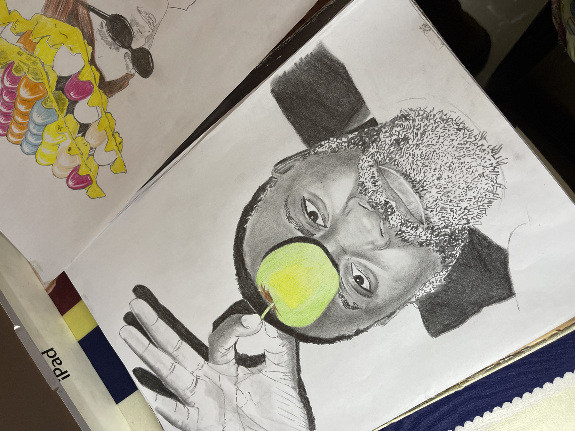 A sketchbook is open, displaying two drawings. The drawing on the right page depicts a man with a full beard, drawn in grayscale. He is holding a green and yellow apple in front of his face. The man has a dark complexion and is wearing a collared shirt or jacket. The drawing is primarily done in pencil, with shading to create depth and dimension. The left page has a colored drawing of another person. The person is wearing sunglasses and has a garland of yellow, white, pink, and orange objects. The background of both drawings is blank white paper. The pages rest on a surface with a striped and patterned edge. Jos, Nigeria is not discernible from the image.