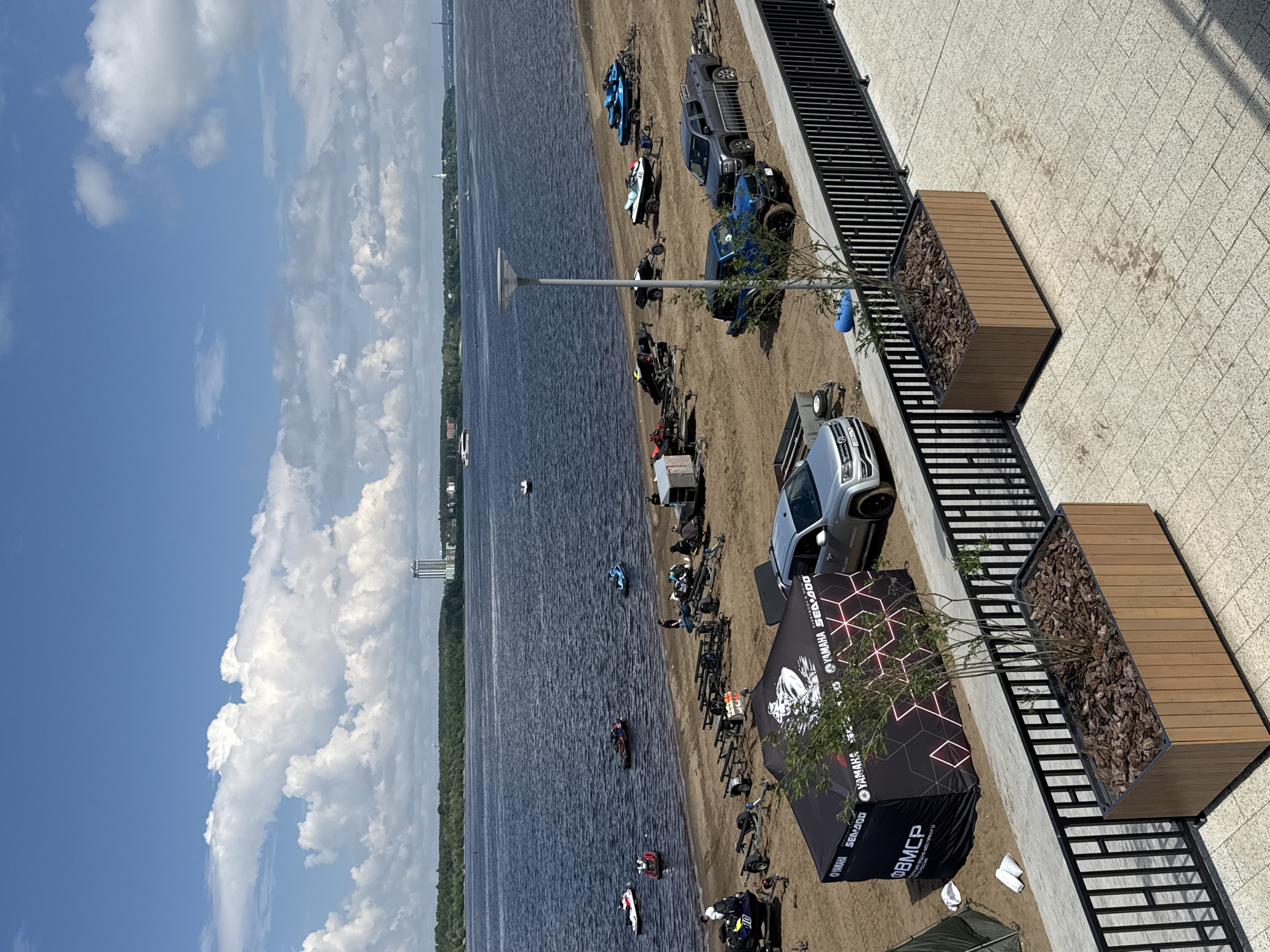 This is an aerial, eye-level outdoor photograph taken on a sunny day, showcasing a scene along the Kama River embankment in the city of Perm, Russia. The foreground features a modern paved area with a black metal railing and two large wooden planters containing small trees and mulch.
Beyond the railing, a sandy embankment is visible, populated with numerous jet skis, some on trailers and some in the water. Several vehicles, including a gray pickup truck, a blue SUV, and a silver SUV, are parked on the sand, some hitched to trailers for the jet skis. A large black and red tent with "YAMAHA SEA-DOO" branding is erected on the sand, likely for an event. A few individuals are scattered on the embankment, appearing to be preparing or managing the jet skis.
The middle ground is dominated by the wide Kama River, with several jet skis actively being used on the water. In the distance, the opposite bank of the river is lined with a dense forest, and further back, a tall, modern building stands prominently against the blue sky. The sky itself is a vibrant blue, punctuated by large, fluffy white clouds, indicative of fair weather. The overall impression is of a recreational area on the riverbank, likely set up for a water sports event.
