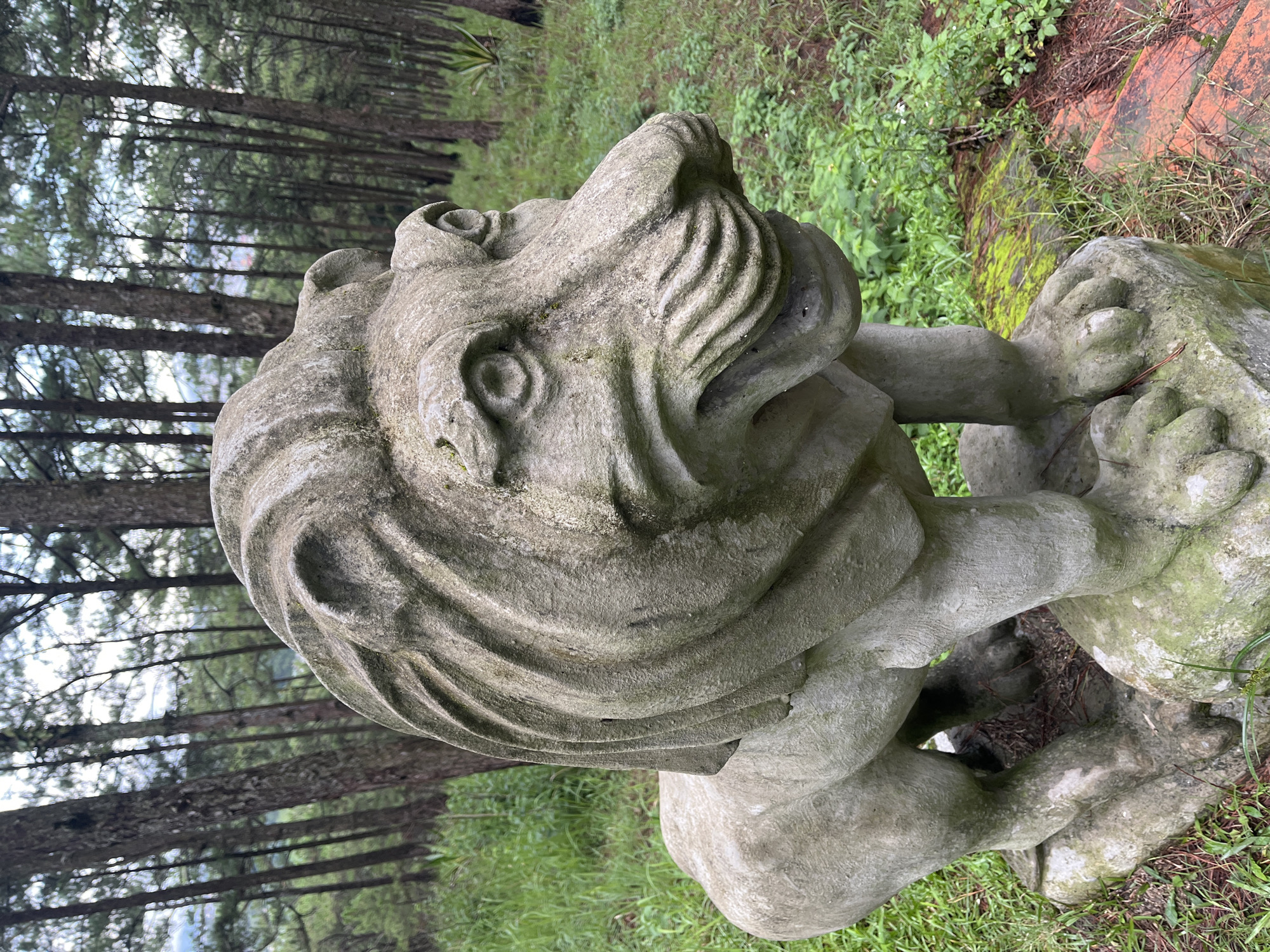 A stone lion statue stands in a wooded area, likely a park or cemetery, with tall pine trees in the background. The statue, weathered and moss-covered in places, depicts a lion in a crouching stance, facing to the right. Its mane is stylized with carved waves, and its facial features, including wide eyes and a snarling mouth, are distinctly rendered. The statue is positioned on a rough-hewn stone base amidst green grass and fallen pine needles. A hint of orange-red brickwork is visible to the right of the statue. This scene is located in Cam Ly, Đà Lạt, Vietnam.