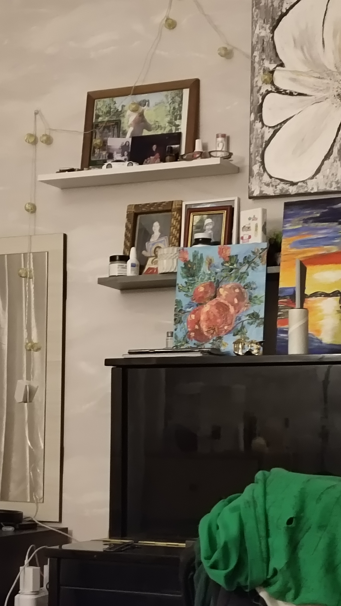 This media file depicts a decorated interior space in Chaikovsky, Russia, likely a living room or bedroom. The main focus is on a light-colored wall adorned with multiple framed pictures, paintings, and decorative items arranged on two floating shelves.
Across the top of the wall, string lights with small, spherical yellow/gold bulbs are draped, suggesting an evening setting or a cozy ambiance.
The upper white floating shelf holds several framed photographs. The most prominent is a large rectangular frame containing a picture of a person, possibly a child, outdoors in a green, natural setting. Below this, smaller framed photos, a white toy car, and a pair of sunglasses are displayed.
The lower, gray floating shelf features two more framed photographs; one in a woven-textured frame and another in a brown and gold frame. Beside them are a small white bottle, a small white jar, and a small religious icon or figurine.
Artworks are a significant feature. To the right, a large painting with a white floral design on a gray and black textured background hangs on the wall. Leaning on the lower shelf is a vibrant painting depicting red-orange round fruits, possibly pomegranates or apples, with green leaves on a light blue background. To its right, another painting portrays a colorful sunset or sunrise over water, using bold strokes of orange, yellow, and blue. A white cylindrical object, possibly a rolled paper or a candle, stands in front of this painting.
In the foreground, a large, dark, reflective piece of furniture, resembling a television or a cabinet, occupies the lower right portion of the frame. A bright green crumpled garment or blanket with a visible hole rests on its surface. To the far left, a tall framed mirror or reflective artwork is partially visible, with power cables and a white charger near its base.
The arrangement of personal photos and decorative elements contributes to a lived-in and personalized atmosphere. No people are actively present in the scene, and no specific activity is taking place. There is no visible text in the image.