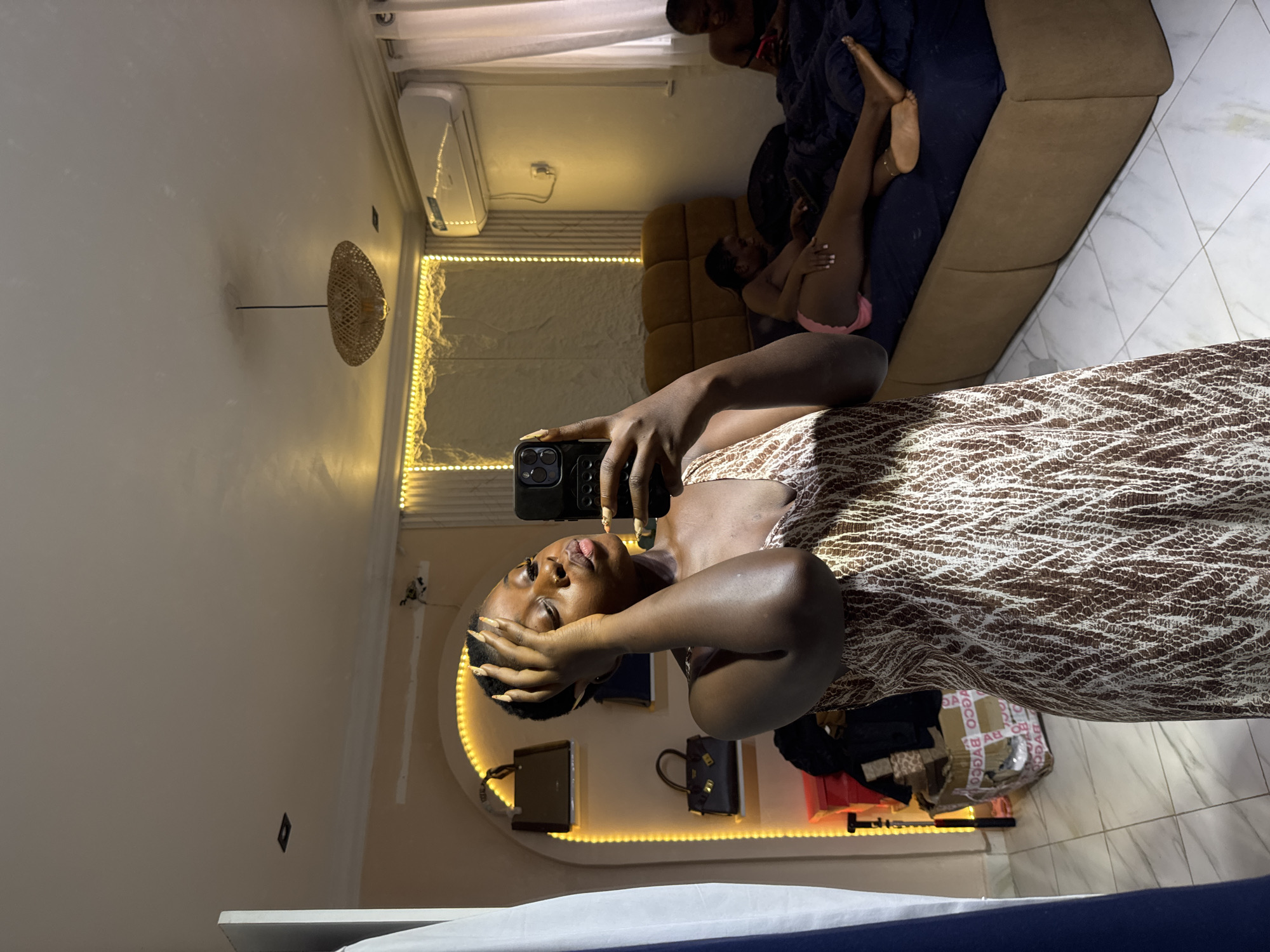 The image depicts an indoor scene, likely a bedroom, bathed in warm, artificial light. In the foreground, a young Black woman is taking a selfie in a mirror. She is wearing a sleeveless, patterned dress and has her hand on her forehead. Her lips are slightly pursed, and she is looking at her phone.
In the background, two other individuals are visible on a bed. One is lying down, facing away from the camera, while the other is sitting up, looking at their phone. The room features a bed with dark blue bedding, a headboard, and a bedside table. Decorative string lights are mounted on the wall behind the bed, casting a warm glow. An air conditioning unit is mounted on the wall above the bed. To the left of the foreground subject, there are shelves with handbags and a stack of boxes. The floor is tiled. The overall impression is of a casual, domestic setting.
While the image is set in a bedroom, there is no specific visual information to confirm it is located in Abuja, Nigeria.