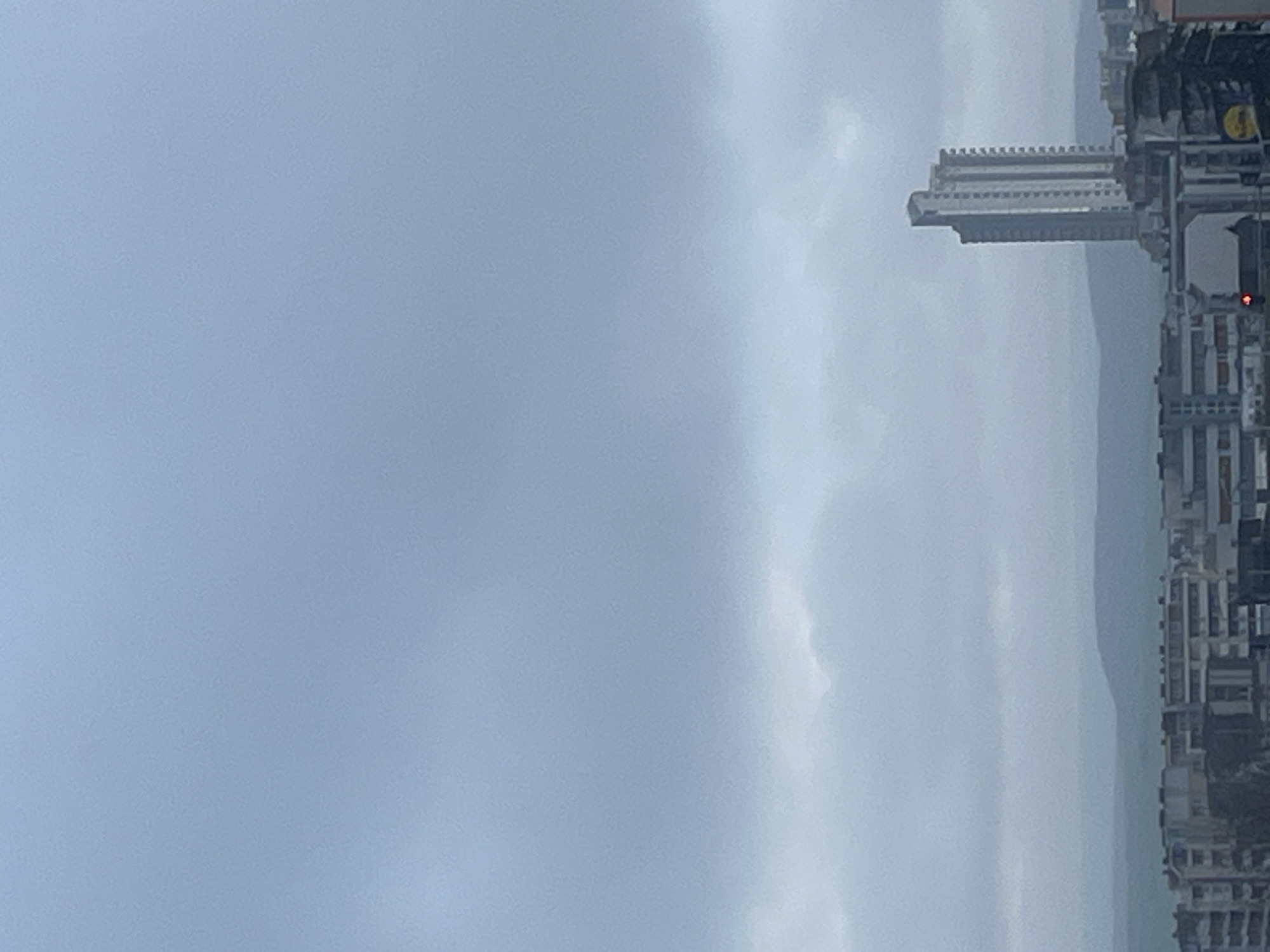 The image shows a cloudy cityscape in Burgas, Bulgaria. Tall buildings line the right side of the frame. To the left of the buildings is a stretch of water. Most of the frame is filled with a gray, overcast sky.