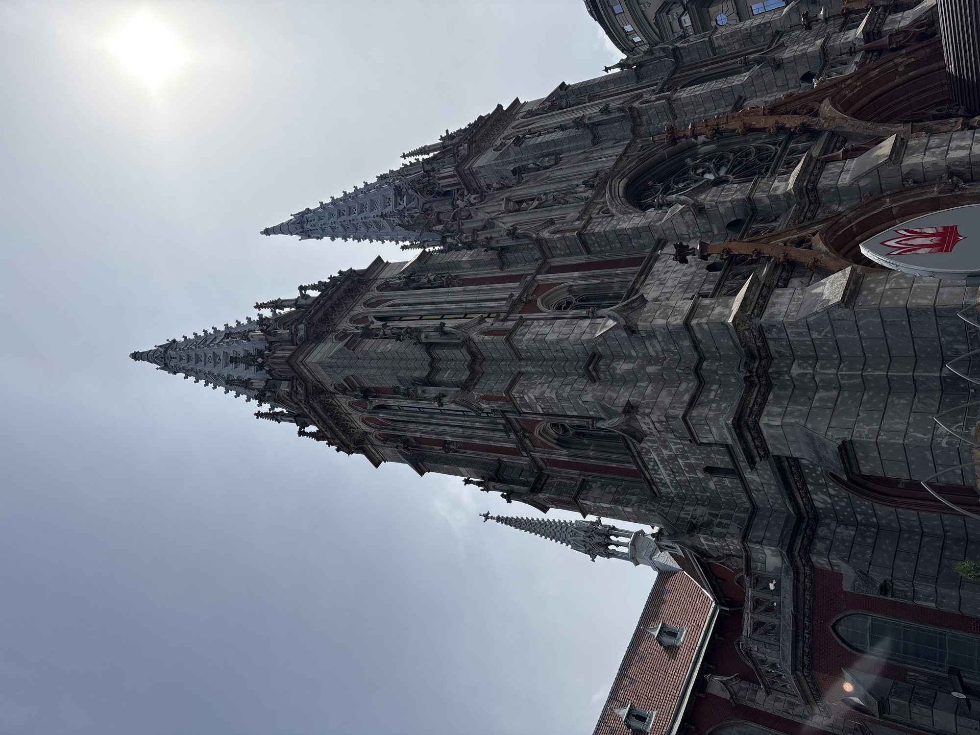The image captures St. Nicholas Roman Catholic Cathedral in Kyiv, Ukraine, viewed from below at an oblique angle. The cathedral's gray Gothic-style architecture with red brick accents rises prominently against a bright, overcast sky. Two spires are visible, one slightly further away. A portion of the adjacent building with a red tile roof is visible in the lower right corner. A sign with a white and red crest is mounted to the building near the base of the frame. The sun is high in the sky. No people are visible in the frame.