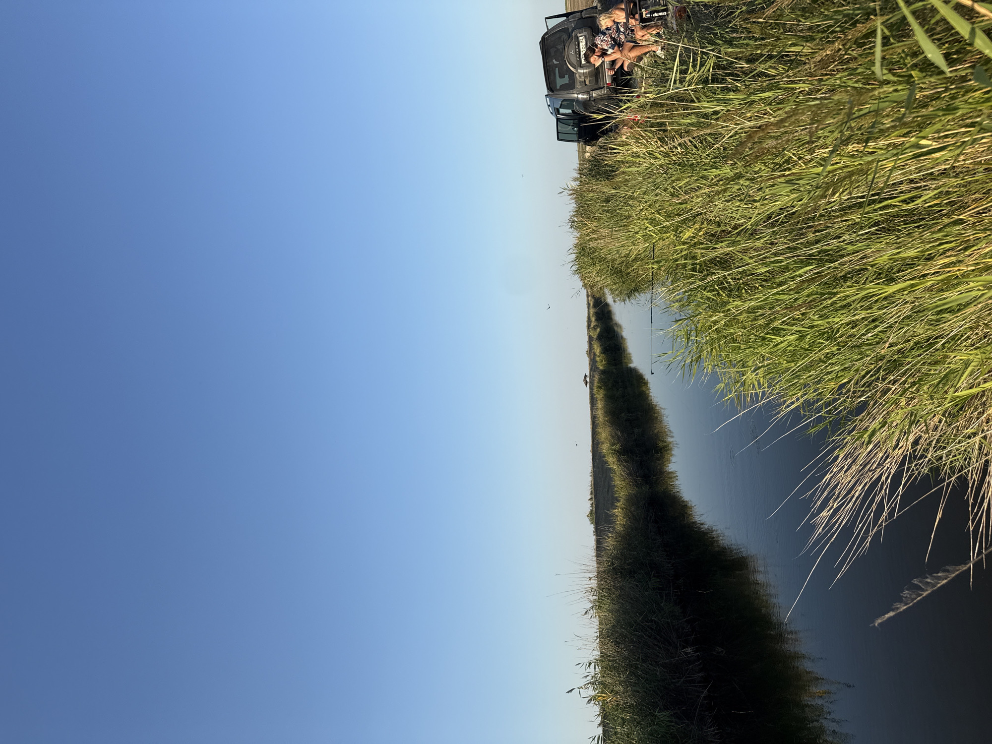 The image depicts a canal lined with tall grass and reeds, reflecting the clear blue sky and a few distant birds. On the right bank, a dark-colored SUV is parked with its rear door open. Two individuals are seated in camping chairs next to the vehicle, with one appearing to be a woman with blonde hair and the other a person wearing a patterned shirt. The car has a spare tire mounted on the back and a license plate that reads "10C BK". Fishing rods are visible in the canal, suggesting recreational activity. The environment is natural and somewhat remote, with a wide expanse of sky and vegetation. The scene appears to be in a rural or natural setting, possibly near the city of Некрасовское сельское поселение, Ukraine, given the provided context.