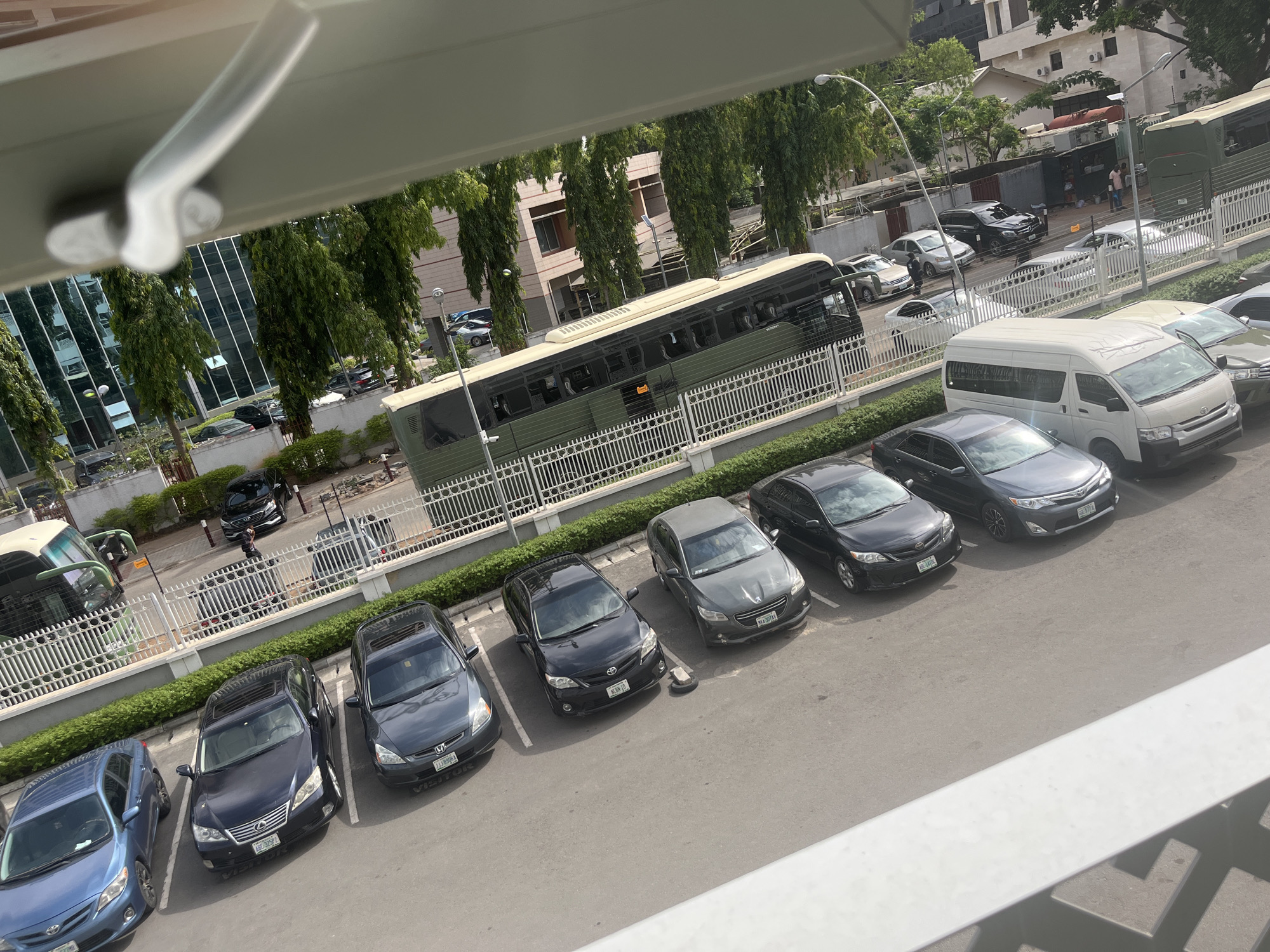 The image captures an elevated view of a bustling urban scene, likely a street and adjacent parking areas, under bright daylight. In the foreground, partially obscured by a white railing or window frame, is a parking lot filled with numerous parked cars. These include several dark-colored sedans, such as a blue Toyota Corolla, a black Lexus, a dark grey Honda Accord, a black Toyota sedan, a grey Peugeot, and another grey Toyota Camry. Many of these vehicles display Nigerian license plates, with codes like "ABC-375-FL", "NSR-17", "MA-007-AV", "11385GA", "BGD-410HG", and "SGE-377XX" visible. One Honda Accord also has "VISITOR" printed on its plate holder. A white Toyota Hiace van is also parked prominently in this section.
Beyond the parking lot, separated by a white picket fence and a green hedge, is a street. A long, olive-green and beige bus with tinted windows is visible on this street, appearing to be parked or moving slowly. Further down the street, more vehicles are parked or in transit, including several SUVs and sedans.
The background features various multi-story buildings, some with modern glass facades reflecting the sky and surrounding trees, and others with more traditional concrete or brick construction. The area is lined with tall, slender trees featuring dense green foliage, characteristic of tropical urban environments. Streetlights are also visible. A few individuals can be seen near the vehicles and fences, suggesting pedestrian activity. The high sun angle and bright illumination indicate the time of day is likely midday or early afternoon, with clear weather. The overall scene depicts a typical commercial or residential streetscape in a Nigerian city like Abuja.