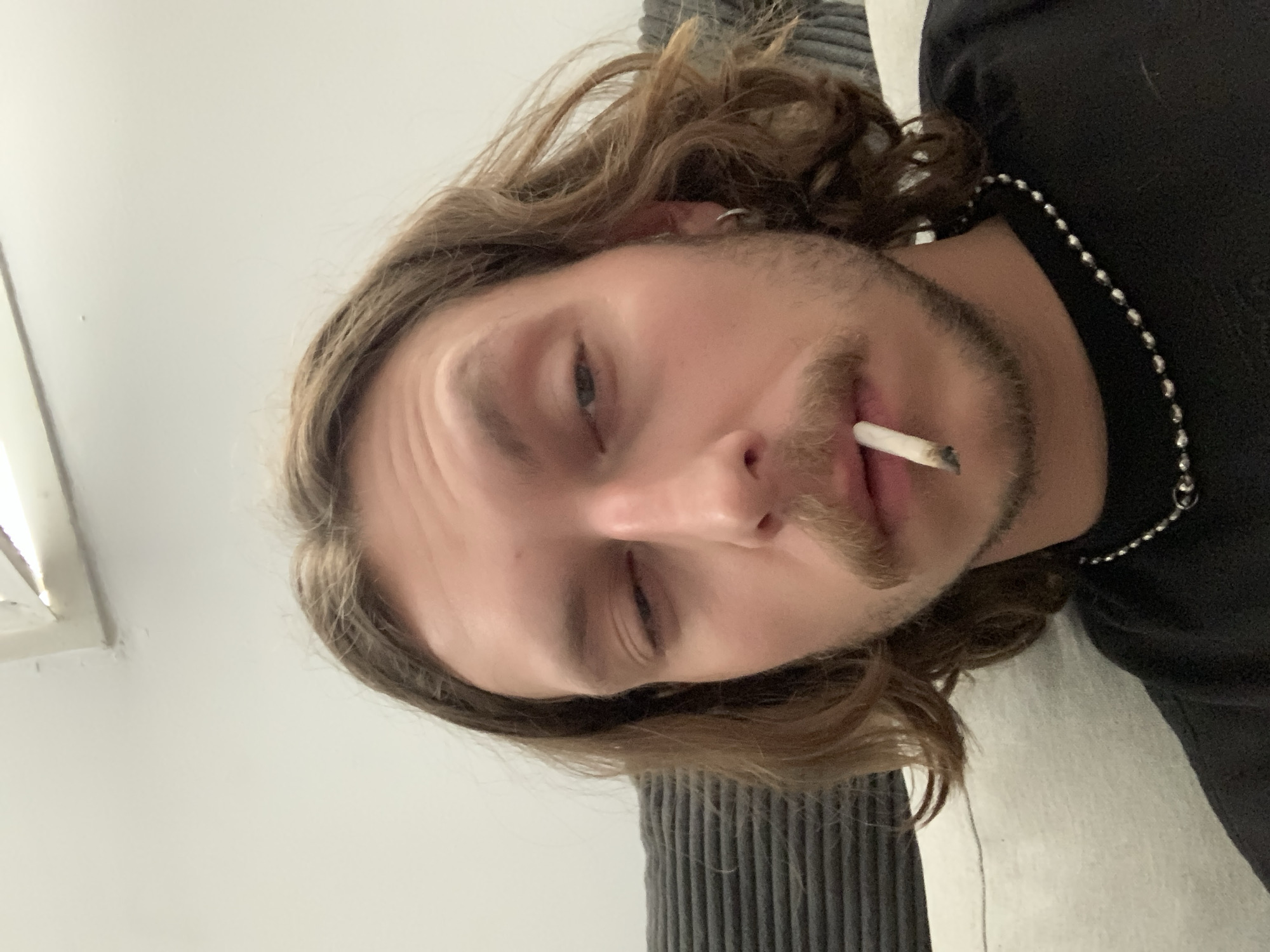A man with long brown hair, a slight mustache and stubble, and a cigarette in his mouth is lying down. He is wearing a black shirt and a silver necklace. His left eye is partially closed. He appears to be indoors. The location is unknown and cannot be determined from the image.