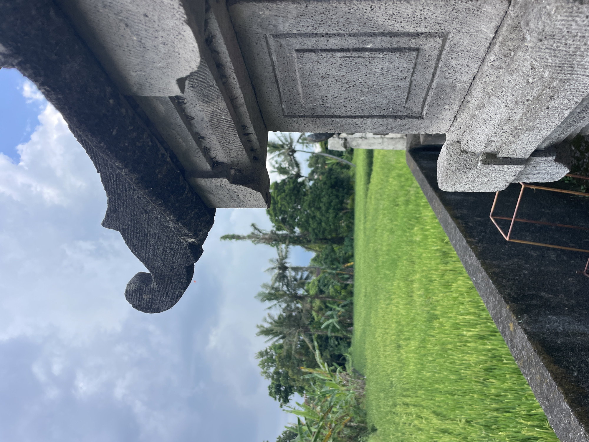 The image shows a view from under a stone structure, possibly a temple or monument, in an unknown location in Unknown. The structure is made of grey, carved stone with geometric and decorative details. Through the opening, a bright green rice field is visible, stretching towards the horizon. Trees, including palm trees, line the edge of the field. The sky is partly cloudy.