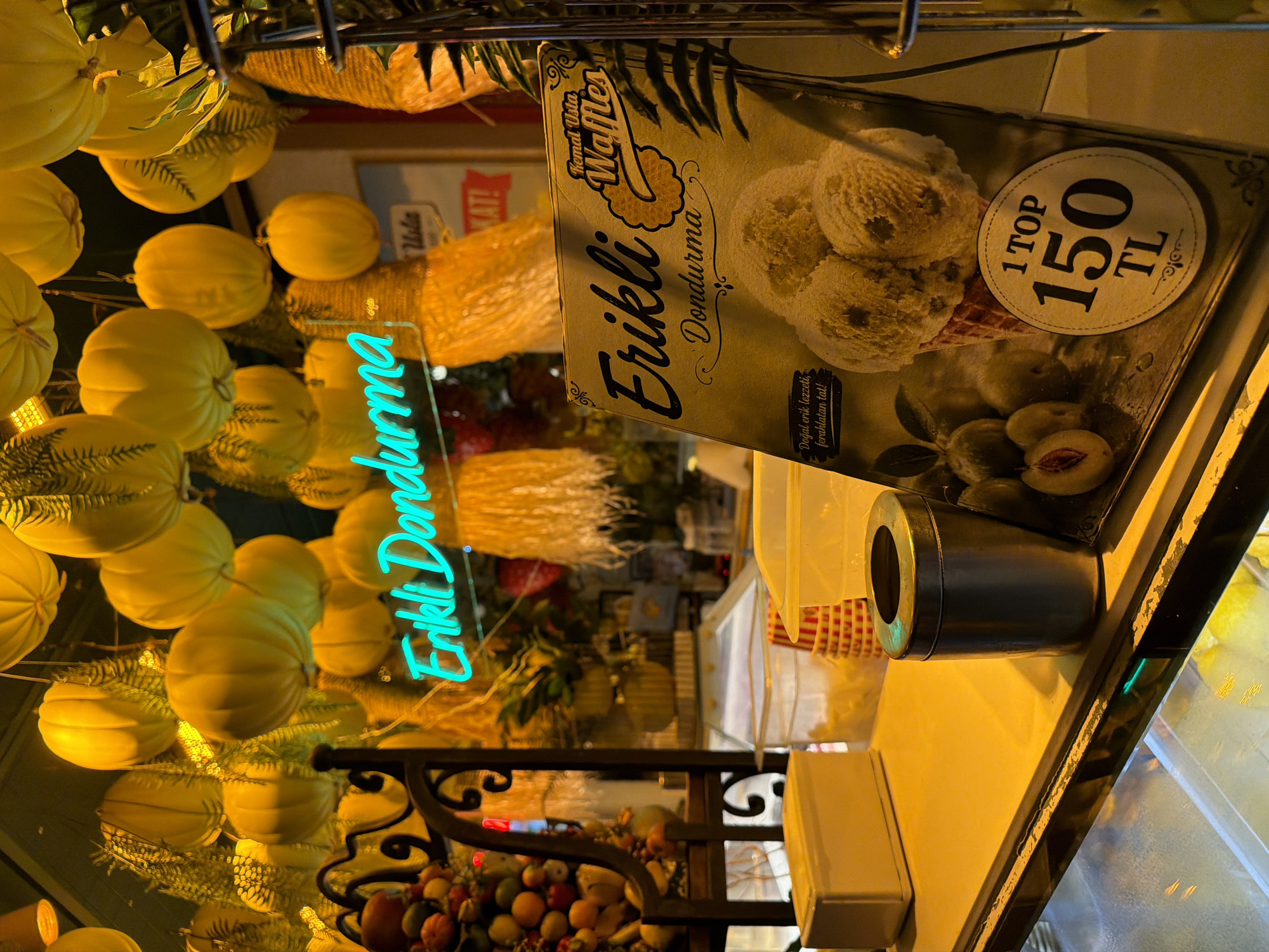 The image captures a close-up view of an ice cream shop, likely in Kadıköy, Turkey, given the signage. Dominating the foreground is a menu or display card featuring a cone piled high with three scoops of ice cream, surrounded by images of plums. Text on the card reads "Erikli Dondurma" (Plum Ice Cream) and "Kemal Usta Waffles," indicating the shop's offerings and possibly the brand. Below the ice cream image, a price of "1 TOP 150 TL" is prominently displayed, suggesting the cost for one scoop.
The background is dimly lit, with numerous round, light-colored decorations hanging from the ceiling, resembling pumpkins or gourds. A bright neon sign in a turquoise color spells out "Erikli Dondurma" across the upper portion of the image, adding to the vibrant atmosphere. Various other elements, such as decorative fringes and greenery, are also visible, contributing to the overall ambiance.
On the counter in the foreground, a yellow rectangular box sits to the left, possibly containing supplies or packaging. Next to it, a metal trash receptacle and some patterned cone cups are visible. The overall impression is one of a cozy, perhaps festive, establishment offering sweet treats. The lighting is warm and inviting, casting a yellow glow over the scene.