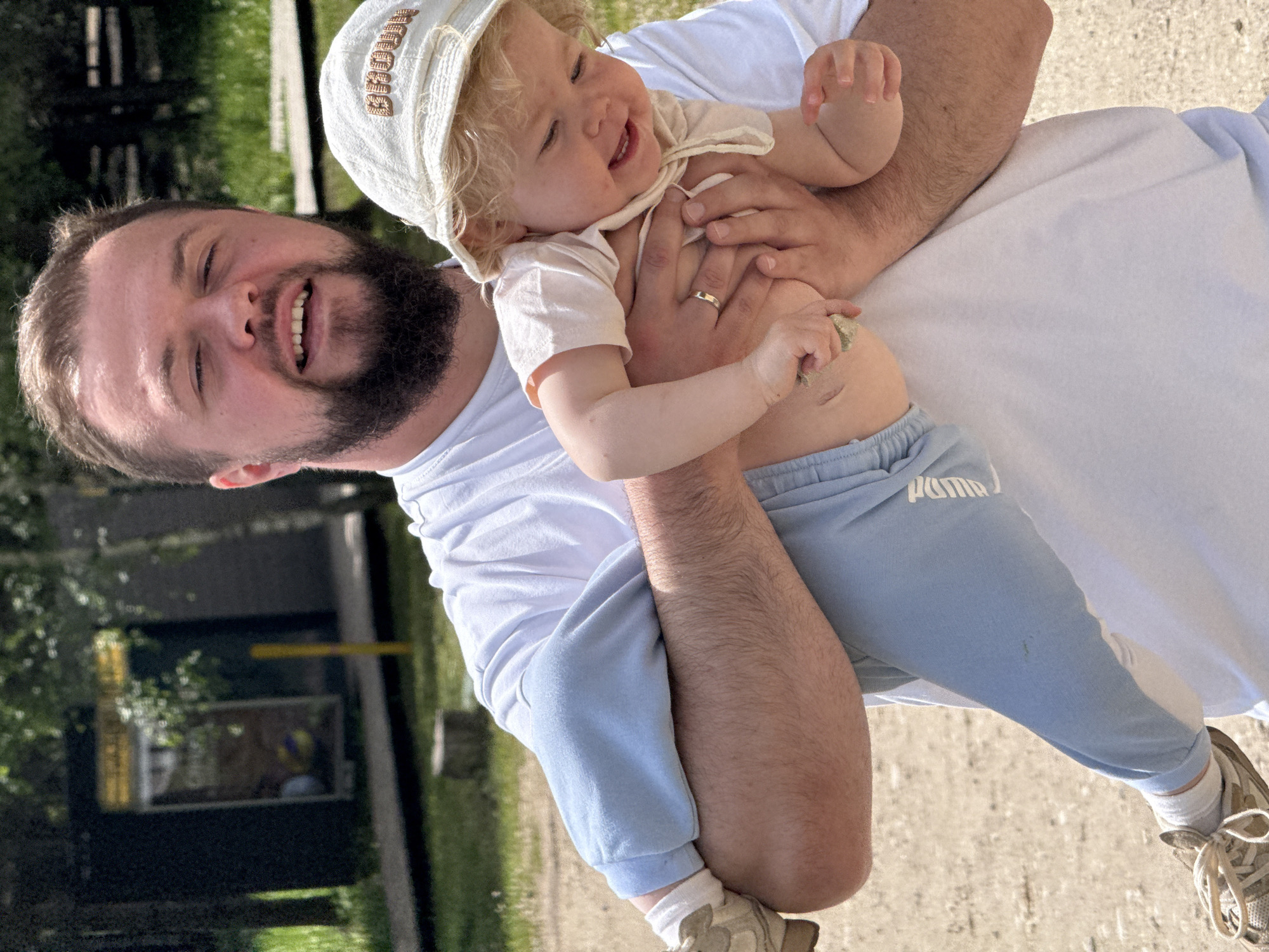 An adult man holds a baby outdoors in a joyful moment. The man, with a beard and short dark hair, wears a white t-shirt, light blue sweatpants, and light-colored sneakers. A wedding band is visible on his left ring finger as he smiles widely, looking down at the baby.
The baby, with blonde curly hair, wears a light-colored bucket hat with "BROWN" embroidered on it, a light short-sleeved top, and light blue sweatpants. "PUMA" is visible on one leg of the baby's sweatpants. The baby is also smiling broadly, looking towards the man, suggesting happiness and engagement.
The scene is set outdoors on a sunny day, likely in a park or recreational area in Dobrograd, Russia. Green trees and foliage are visible in the background, with a light-colored paved ground beneath them. The bright lighting indicates daytime. The overall emotion conveyed is one of warmth, affection, and happiness between the two individuals.