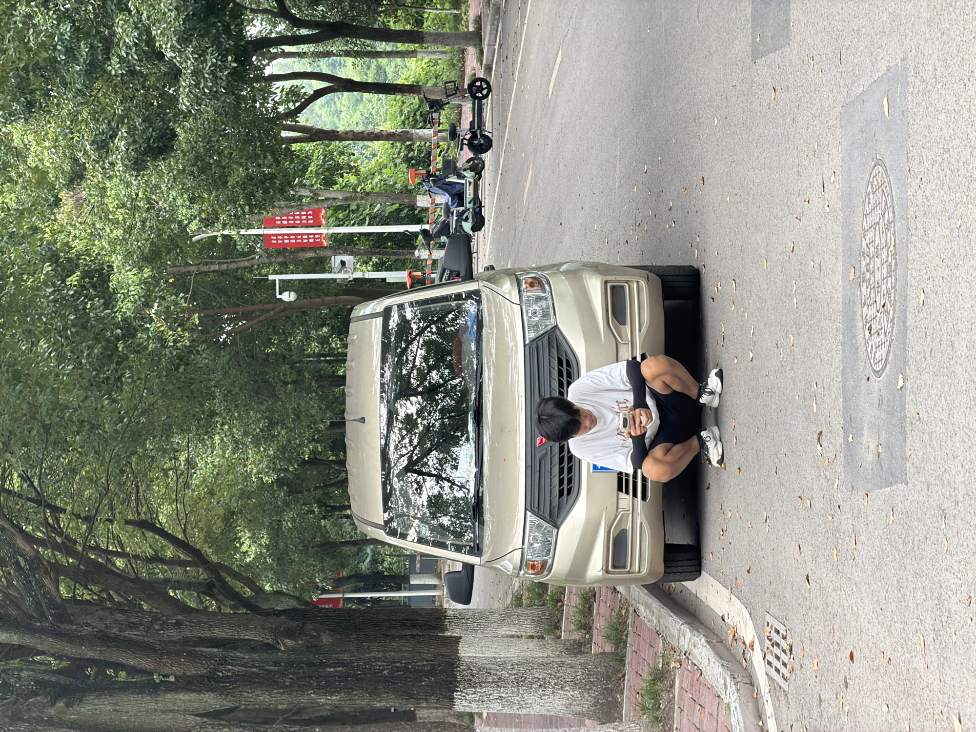 In the city of Jiangjin, China, a young East Asian male is crouched on an asphalt road, looking at his phone. He is wearing a white t-shirt, black shorts, and white sneakers. He is positioned in front of a gold minivan. The minivan is parked on the side of the road, facing forward. The road is lined with trees on both sides. There are fallen leaves scattered on the road and sidewalk. In the background, several electric scooters are parked. Red banners with Chinese characters are hanging from a lamppost. The scene appears to be a residential or commercial area, with a drain grate visible on the left side of the road and a manhole cover in the foreground.