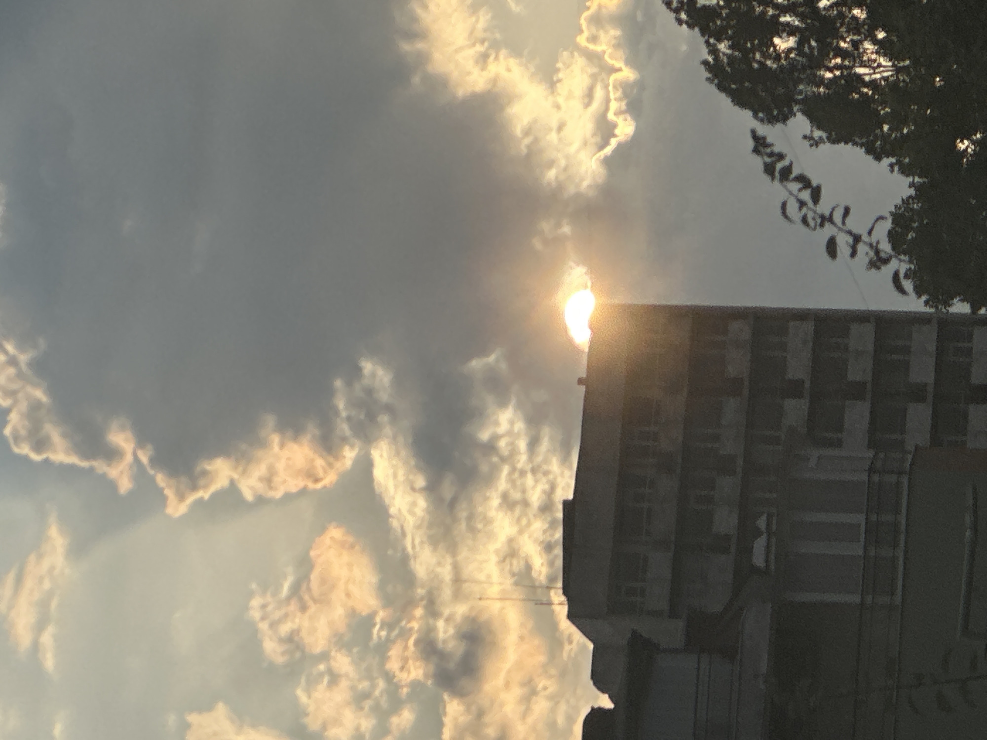 A building is visible in the foreground of the image, partially obscured by trees. The building appears to be a multi-story structure with a modern architectural style. The sky is filled with dramatic clouds, with the sun peeking through them, creating a warm, golden light. The scene captures a moment of natural beauty in an urban setting.