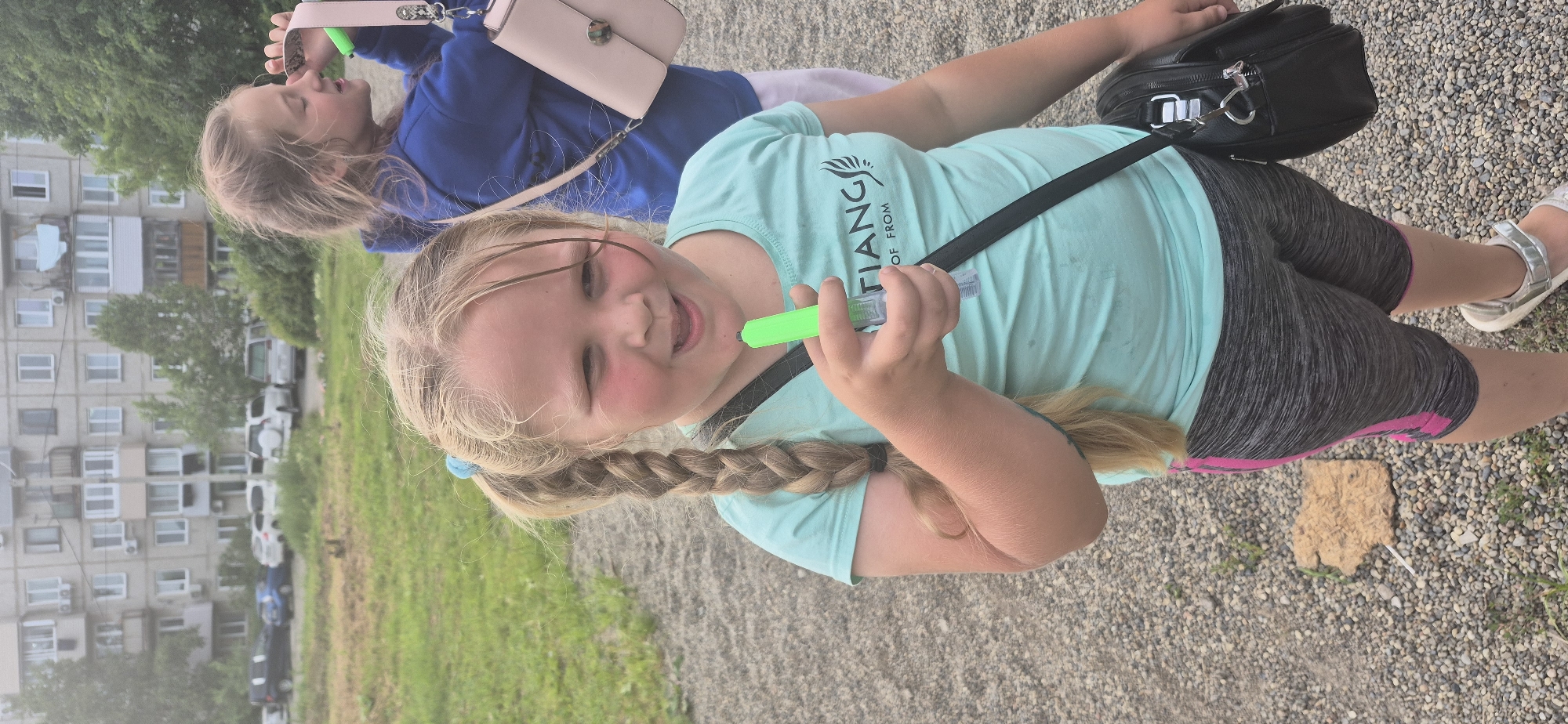 Two young girls are pictured outdoors in a gravel area with grass and buildings in the background. The girl in the foreground is facing the camera, smiling, and holding a green marker. She has blonde hair in a braid and is wearing a light blue t-shirt and grey shorts with pink stripes. She has a black bag slung across her body. The second girl, in the background, is wearing a blue hoodie and is holding a pink purse. She is looking away from the camera. In the background, there are apartment buildings and several parked cars. The lighting suggests an overcast day.