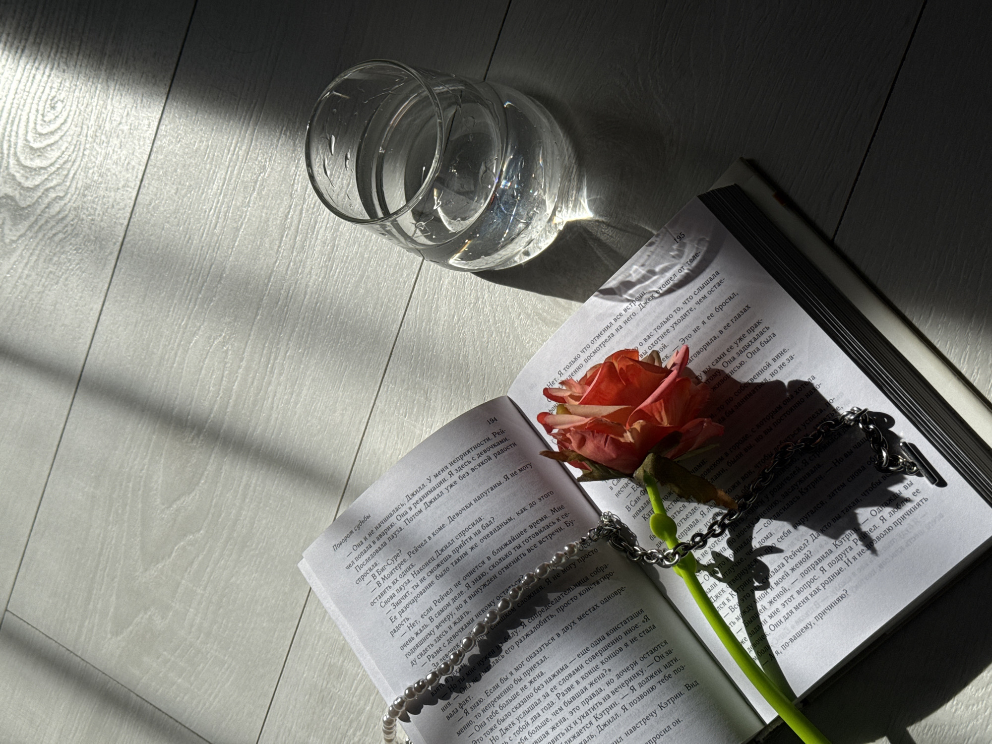 An overhead shot captures an open book, a single rose, a clear glass, and jewelry laid out on a light-colored wooden floor. Distinct, diagonal shadows are cast across the scene, indicating strong, direct sunlight, likely from a window, suggesting it's either morning or late afternoon.
The book, open to pages 194 and 195, features text in Russian. The title "Поворот судьбы" (Turn of Fate) is visible on page 194. The visible narrative discusses characters named Jill (Джилл), Rachel (Рейчел), Jack (Джек), and Katherine (Кэтрин), touching upon events such as an accident involving Rachel and a discussion about a cancelled ball.
A pink-orange rose rests across the spine and left page of the book. A white pearl necklace and a silver chain with a toggle clasp are draped over the right page of the open book and parts of the rose. A clear glass, possibly a stemless wine glass or water glass, is positioned to the upper right, reflecting and refracting the sunlight onto the wooden surface. The floor planks exhibit light wood grain patterns and distinct seams. The arrangement suggests a tranquil, personal moment of reading or reflection.