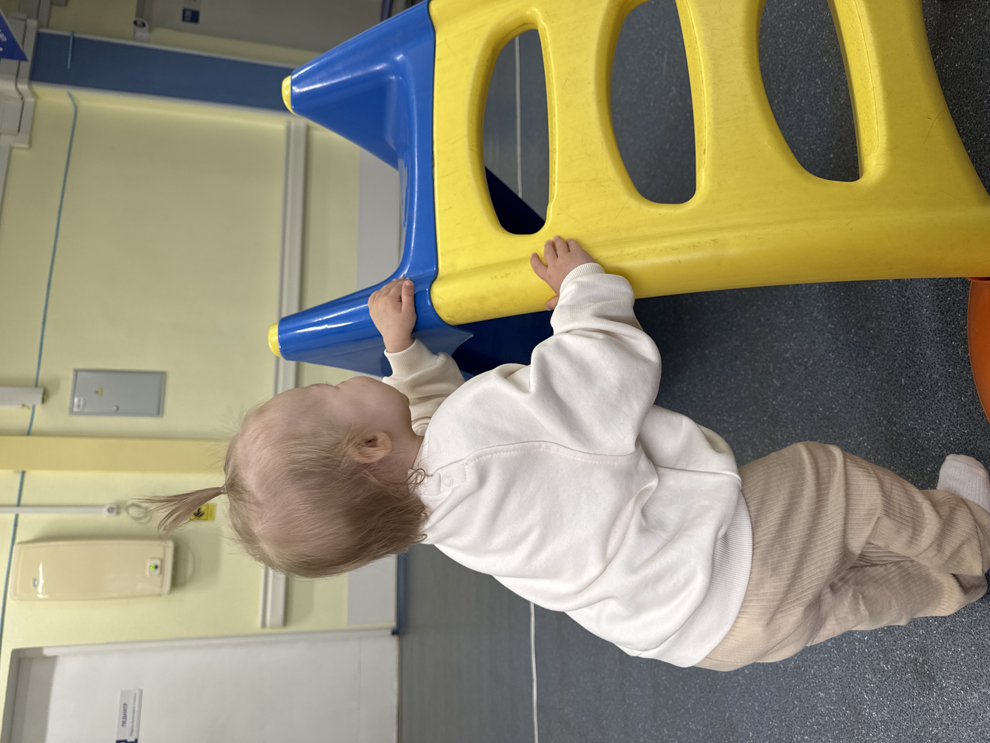 A baby, likely a toddler, with light-colored hair tied in a small ponytail, is depicted standing and engaging with a blue and yellow plastic play structure in an indoor setting. The baby is wearing a long-sleeved, off-white or cream-colored top with buttons on the back of the neck, and light beige corduroy pants, complemented by white socks. Their small hands are gripping the yellow section of the play equipment, which has rounded cut-outs.
The background features light yellow walls with a horizontal blue stripe, suggesting an institutional environment. Various fixtures are mounted on the wall, including a gray electrical box and a cream-colored box with a green indicator light. The floor is a dark gray, possibly linoleum or rubber, showing some scuff marks. A white door is partially visible on the right side of the frame, bearing the partially legible text "ПЕДИАТР," which is Russian for "pediatrician." This detail, combined with the sterile appearance of the room, strongly indicates the location is a children's play area or waiting room within a medical facility, such as a clinic or hospital, in Chaikovsky, Russia. The time of day is not discernable, but the lighting suggests an interior space during operational hours.