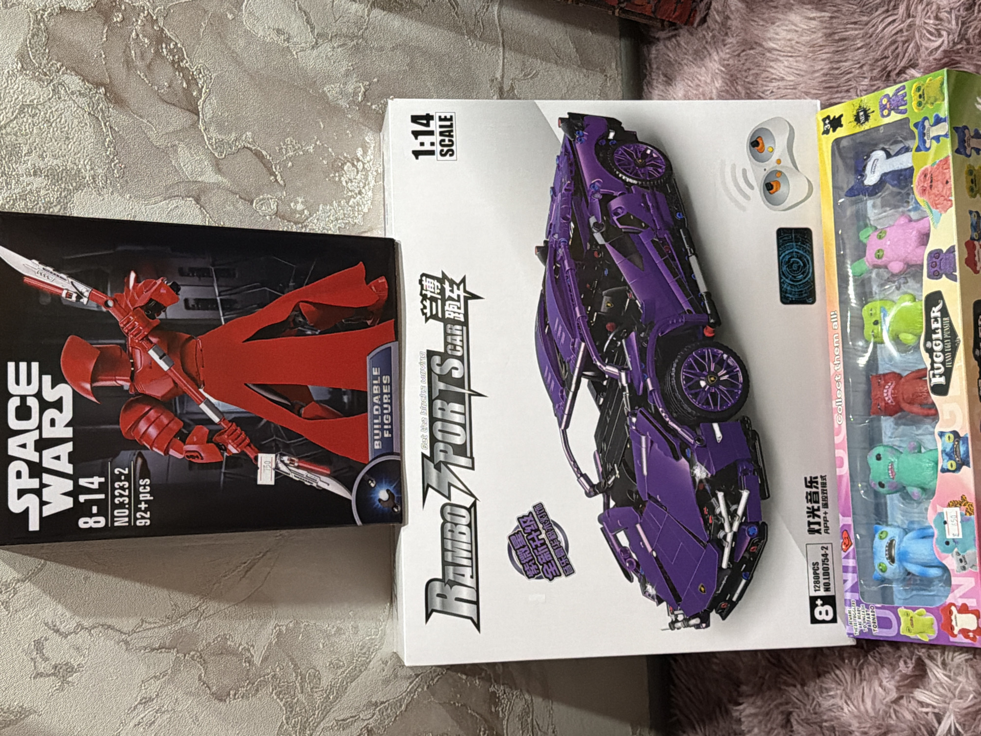 The image shows three boxes of toys or building kits on a surface that has two different textures. On the left is a box labeled "SPACE WARS," with the age range 8-14 years and "92+pcs." The image on the box depicts a red figure with a weapon, presumably a character from the Space Wars theme. To the right of the "SPACE WARS" box is a white box with a purple toy car, labeled "RAMBO SPORTS CAR" with a scale of "1:14." The box also has Chinese characters and indicates "1280PCS" and an age recommendation of "8+". Finally, a box of "FUGGLER" toys is on the far right of the image. The surface beneath the boxes appears to be made up of two different sections: the first is a gray patterned surface, and the second is a fluffy, pink surface. There is no information in the image suggesting the location is Perm, Russia.
