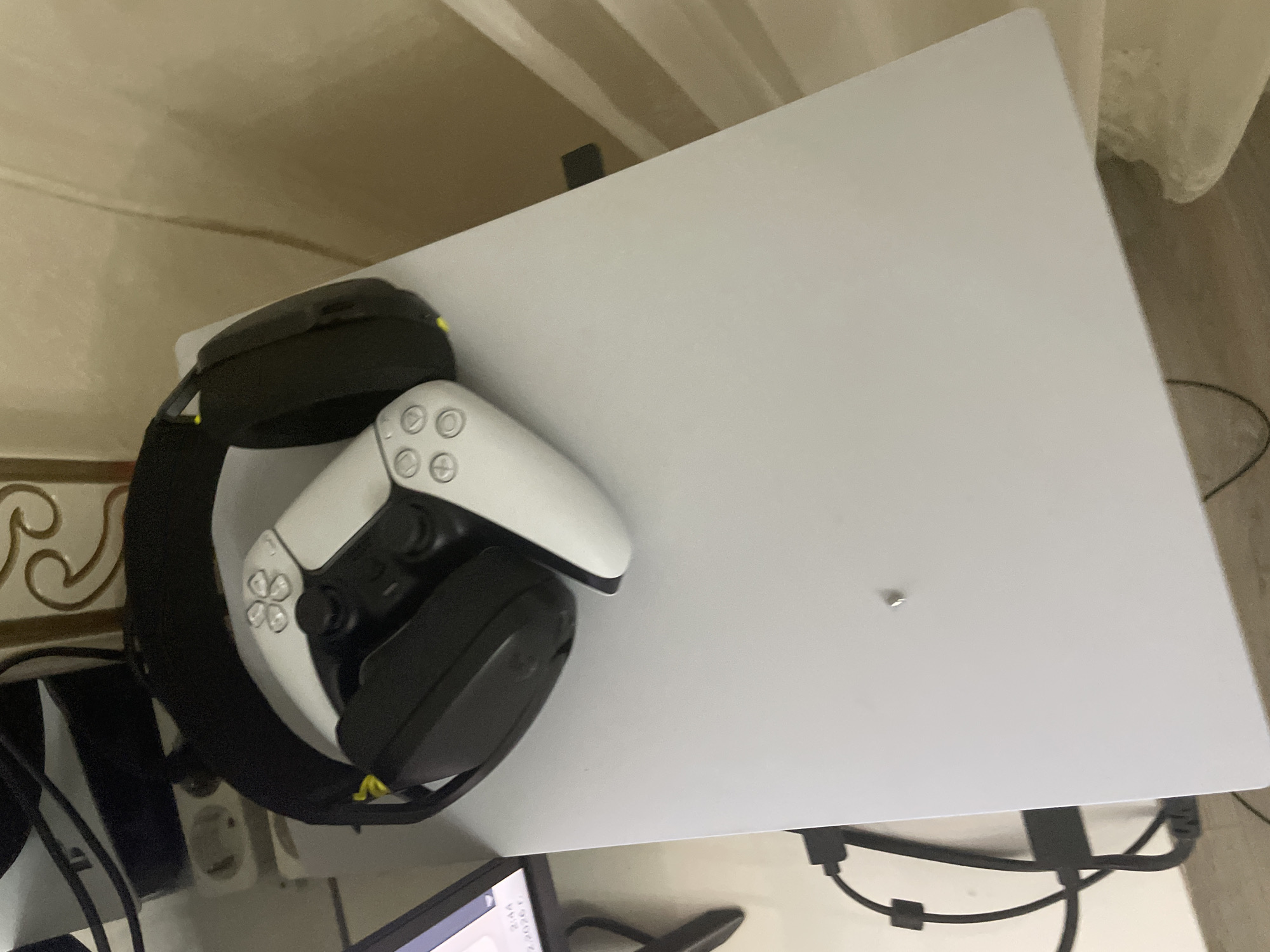 An angled image captures an indoor gaming setup, primarily featuring a white PlayStation 5 console laid horizontally on a surface. Resting on the console's upper white panel are a black over-ear gaming headset, accented with a yellow strap on its left side, and a white DualSense controller. The controller is positioned with its face buttons visible, displaying the standard PlayStation symbols (X, Circle, Square, Triangle) and the PlayStation logo on the touchpad. A small, shiny metallic screw head is visible on the white console panel, typical for a horizontally oriented PS5 stand.
In the background, a light beige or cream-colored wall is visible, featuring a decorative, possibly gold-toned, ornate pattern in the bottom left section of the frame. A light-colored, sheer curtain or fabric hangs in the upper right. Numerous black cables are seen trailing from behind the console, suggesting power and display connections, and further cables are visible towards the bottom of the image. A European-style two-pin power outlet is partially visible on the wall behind the headset. In the very bottom left corner, a portion of a digital screen, possibly from a laptop or monitor, is visible, displaying blurry text and numbers, which appear to include "2:44" and "23 23:26" or similar. The lighting is soft, suggesting an indoor setting. No people are present, and no specific activity is taking place beyond the arrangement of the gaming equipment. The scene lacks specific cues for time of day or weather, but indicates a personal space in Ihtiman, Bulgaria.