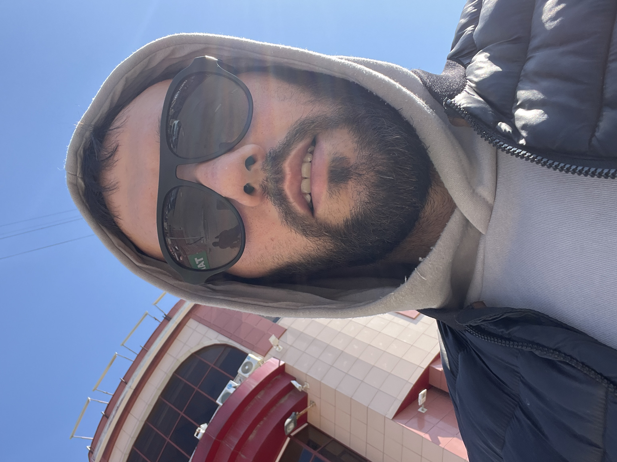 A selfie of a man with dark hair and a beard, wearing a gray hoodie and black sunglasses, standing outside a building on a sunny day. The building appears to be the Ulyanovsk State Pedagogical University, based on the architectural details. The man is smiling slightly. The picture is taken from a low angle, looking up.
