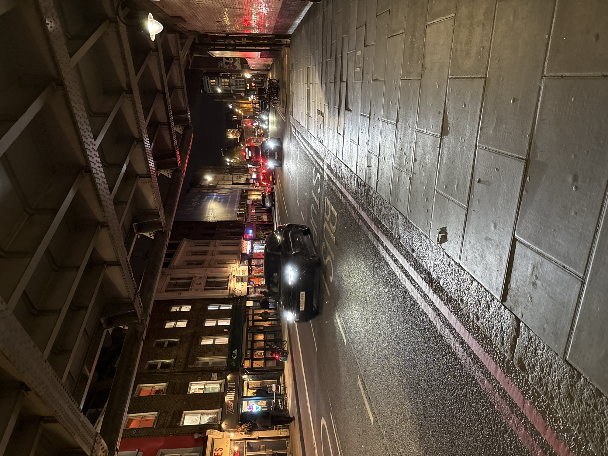 The image shows a street in London, United Kingdom, at night. The street is lined with buildings on one side and a raised railway or road structure on the other. Cars are driving on the wet asphalt road, and the word "BUS" is painted on the street. People are walking and standing on the sidewalks. Streetlights and building lights illuminate the scene.