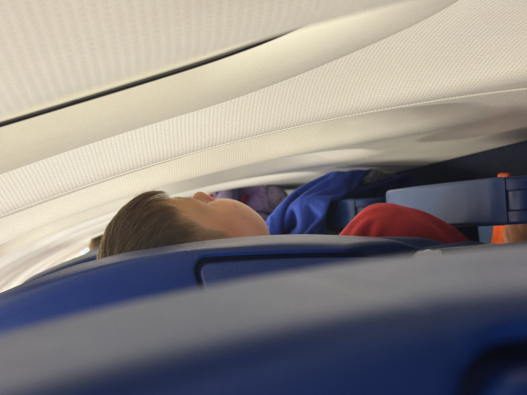 The image depicts a young boy seated on an airplane. He is wearing a red shirt and a blue hoodie, and his face is partially obscured as he looks towards the window. The airplane's interior is visible, with blue seats and a textured white wall. The scene is well-lit, suggesting daylight. There is no indication of the city of Sochi, Russia, in the image.