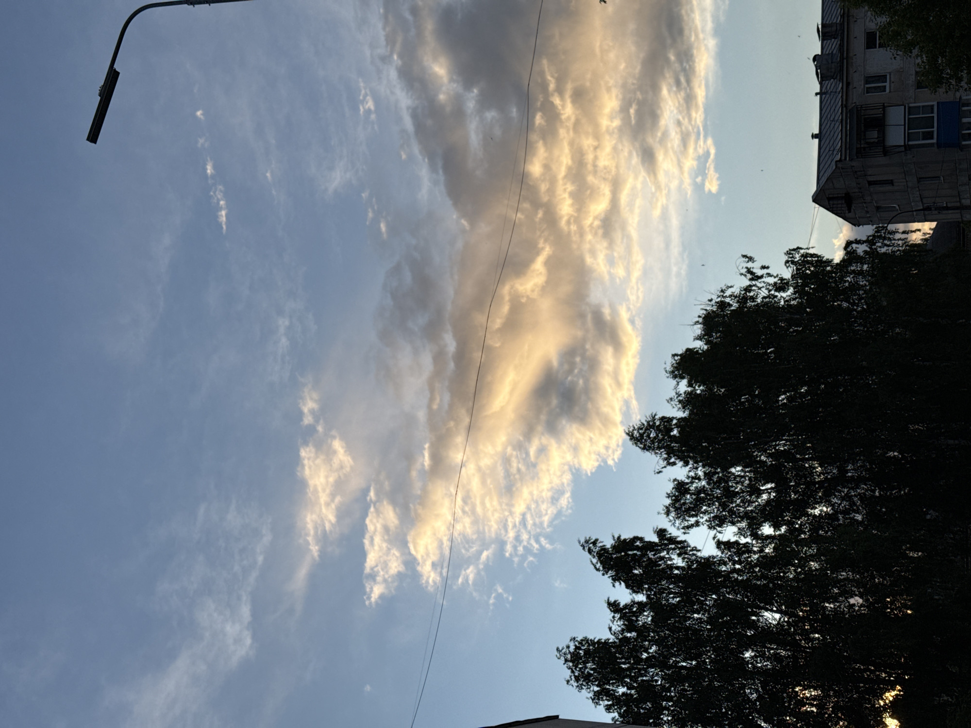 The image depicts a sky scene in Chaikovsky, Russia, captured during daylight hours. A tall, grey, Soviet-era apartment building with multiple windows is partially visible in the upper right. A large, dark tree obscures the lower right corner of the view. A utility pole and wire run across the image. The primary focus is on the sky, filled with clouds illuminated by the sun, with the lower sky a clear blue.