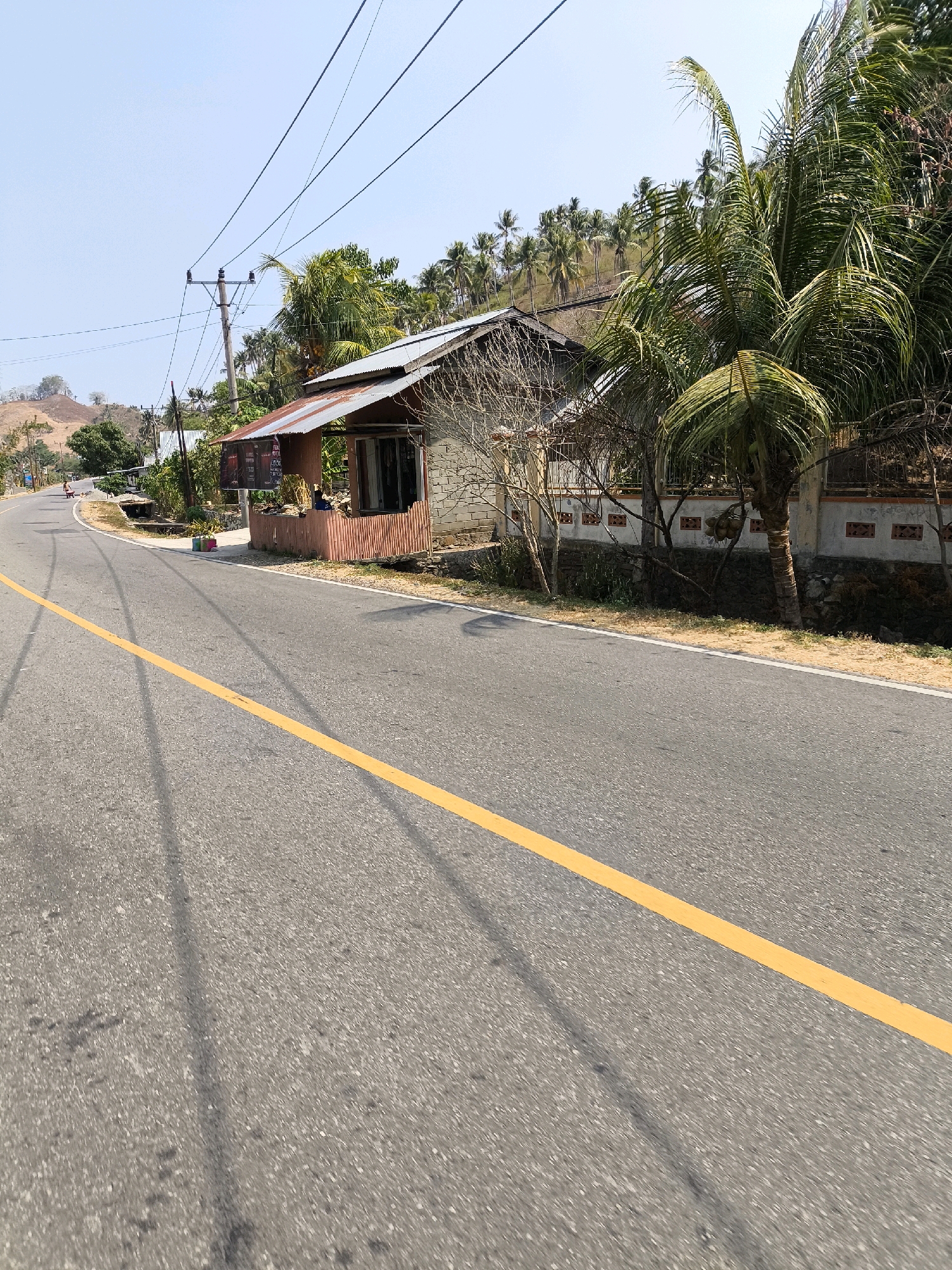 In a sunlit outdoor scene in Unknown, Indonesia, a paved road with a yellow dividing line curves to the left. On the right side of the road, a small building with a corrugated metal roof and a wooden veranda is visible. A sign board with text is mounted on the veranda. A person is sitting on the veranda. Coconut trees and other vegetation are present behind the building. In the distance, hills covered with palm trees and a clear blue sky can be seen. Electrical poles and wires are present throughout the scene.