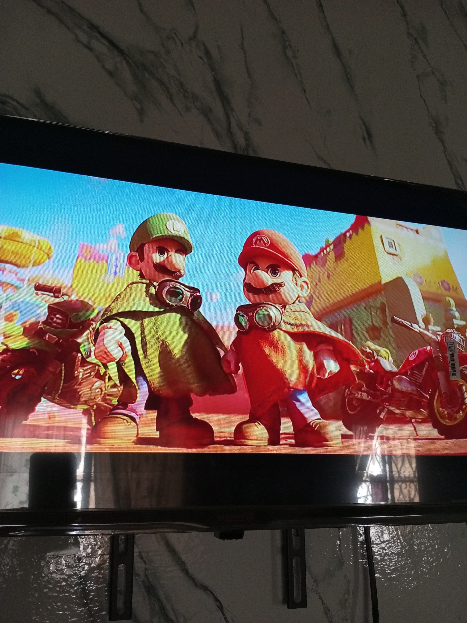 The image shows a television screen displaying an animated scene featuring Mario and Luigi. Both characters are wearing hats, mustaches, goggles and capes, and are standing next to motorcycles. The background depicts a town with buildings and a blue sky. There is no indication of the location being Palo Negro, Venezuela, in the image.