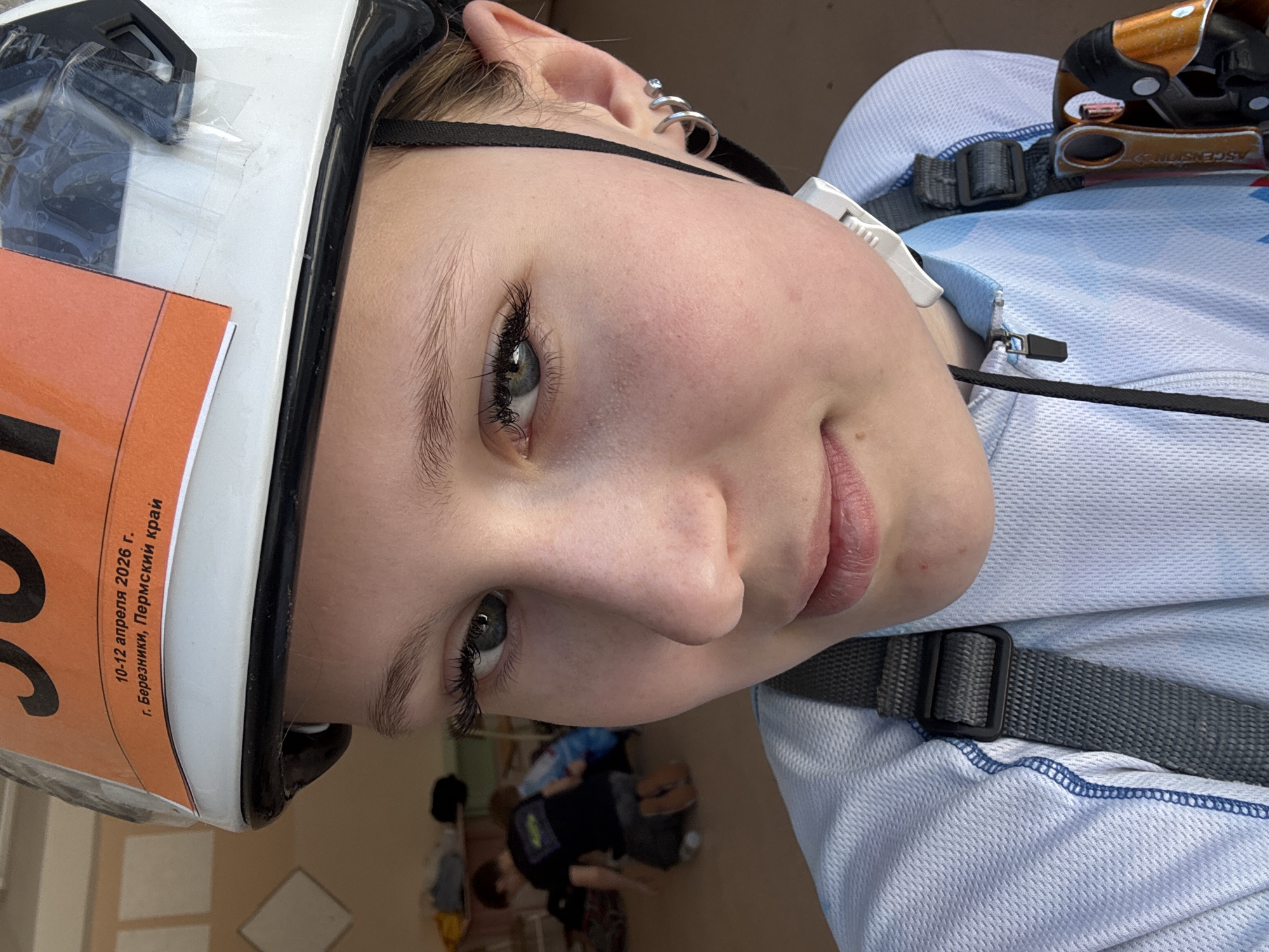 A young woman is shown close-up, wearing a white climbing helmet with an orange sticker reading "10-12 апреля 2026 г. г. Березники, Пермский край" in Cyrillic, which translates to "April 10-12, 2026, Berezhniki, Perm Krai". She has fair skin, gray-blue eyes, and multiple earrings in her left ear. She's wearing a light blue technical shirt and climbing gear, including a chest harness with gray straps and an ascender visible at the top right. A dark cord is also visible around her neck. The background is blurry and indistinct, but an interior location is suggested. While the sticker references Berezhniki, Perm Krai, there is no visual evidence connecting this image to the city of Usolye.