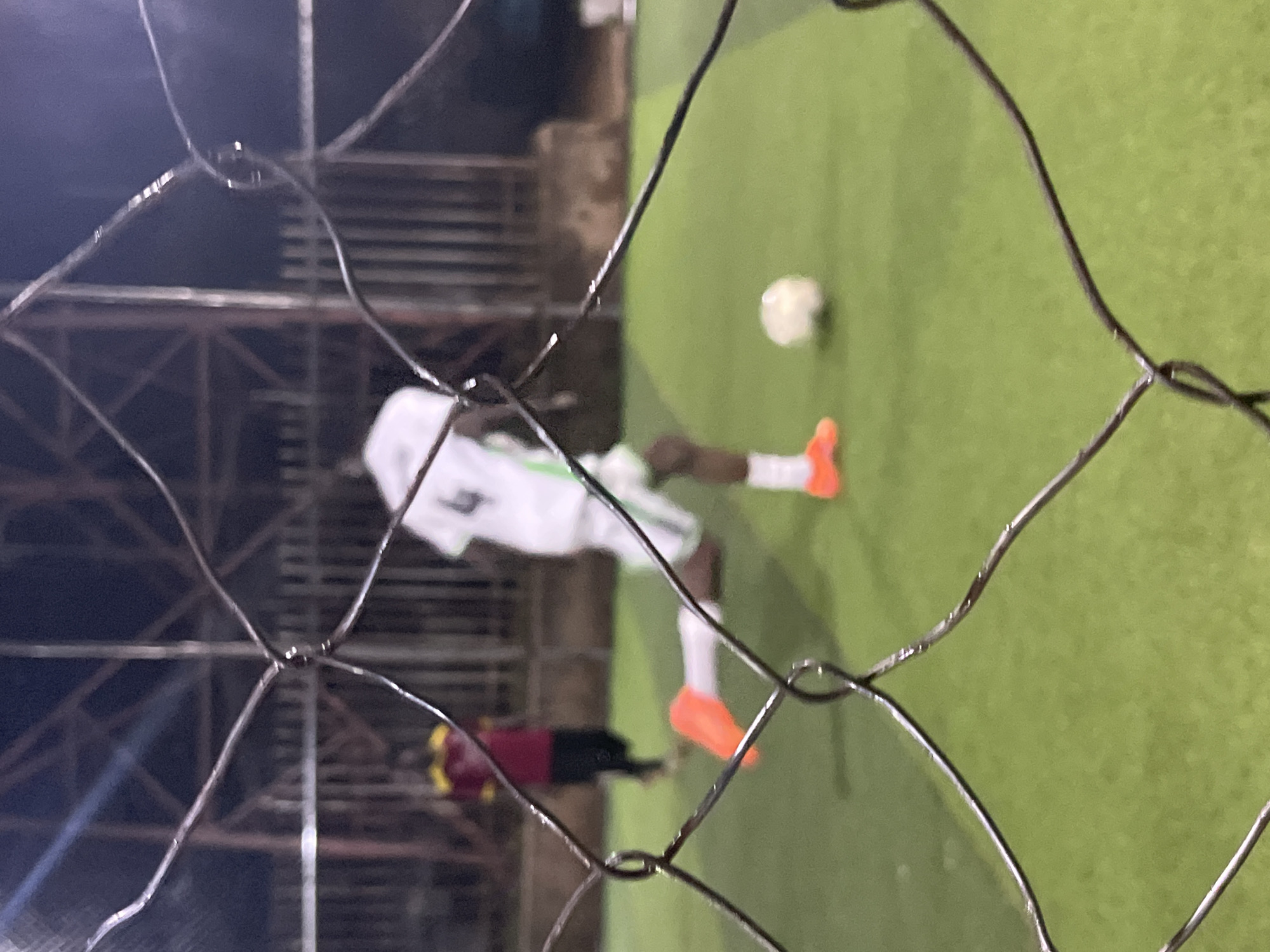 A slightly blurred, nighttime outdoor shot shows a football game being played on an artificial turf field at the University of Jos, Nigeria. A player in a white jersey with green trim and the number 4 on the back is in motion, with their right leg extended. They are wearing bright orange football boots. Another player in a maroon and yellow jersey is visible in the background, to the left. A white football is on the ground between the two players. A wire fence is prominently in the foreground, partially obscuring the view of the field. The lighting appears to be artificial, likely from stadium lights.