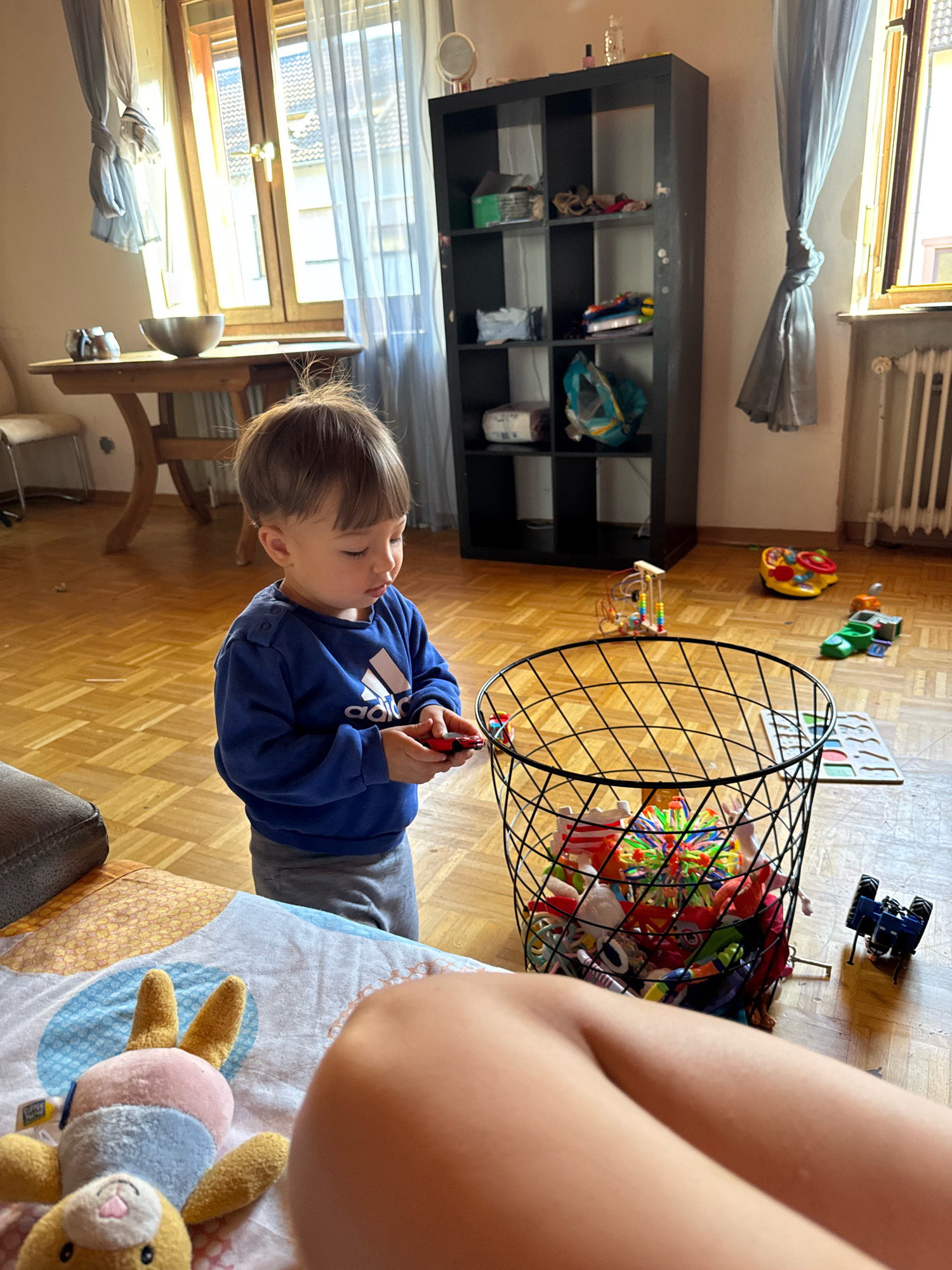 A toddler boy stands in a living room with wooden floors, holding a small red toy car. He is wearing a blue sweatshirt with an Adidas logo and grey sweatpants. In front of him is a black wire basket overflowing with toys, including a colorful spiky ball and various other small items. Behind the basket, several other toys are scattered on the floor, including a yellow and red musical toy, a green toy car, and a blue toy tractor. To the left of the boy, a round wooden table holds a metal bowl and two smaller metal objects. Further in the background, a large black shelving unit is filled with various items. Curtains are drawn across a large window on the left side of the frame, and another window with a radiator is visible on the right. The overall setting appears to be a home in Überherrn, Germany.