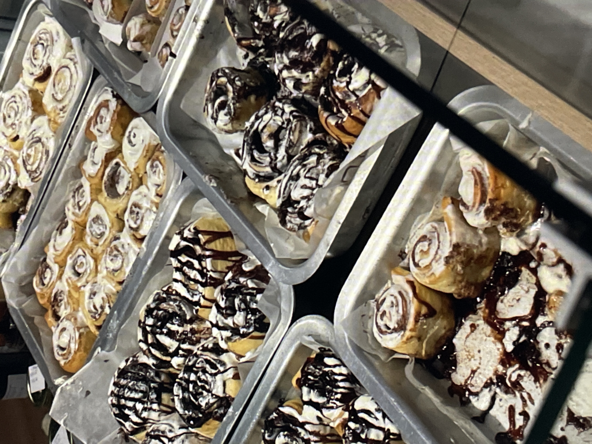 The image displays a multi-tiered commercial presentation of various sweet rolls, likely cinnamon rolls, in multiple rectangular metal trays lined with white parchment paper. Approximately six trays are visible, arranged diagonally and vertically, indicating a display case setting. The rolls are baked to a golden-brown hue and feature distinct toppings: some are covered with a thick white glaze or icing, others are generously drizzled with dark chocolate, and some exhibit a combination of both white icing and dark chocolate drizzle. The rolls are predominantly spiral-shaped. The trays are well-stocked, with some showing minor disturbances in arrangement, crumbs, or residual chocolate and icing on the parchment. The scene is illuminated by warm, artificial lighting. A dark, reflective surface, possibly glass, partially obscures the foreground, creating a diagonal reflection across the lower right portion of the image. No individuals, direct actions, or interactions are visible. The commercial establishment is located in the city of Saint Petersburg, Russia.