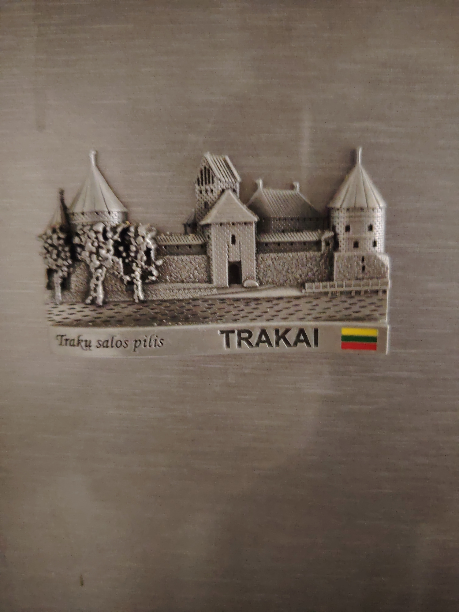 The image displays a silver-colored souvenir magnet featuring a detailed relief of Trakai Island Castle, characterized by multiple fortified towers and buildings with pitched roofs, surrounded by stone walls. Below the castle, the text "Traku salos pilis" is inscribed on the left, identifying the castle's name in Lithuanian, and "TRAKAI" on the right, accompanied by a small rectangular flag with horizontal stripes of yellow, green, and red, representing the national flag of Lithuania. The magnet is presented against a subtly textured, brushed metallic background. No people are present, and no actions are occurring within the scene. The location relevant to this image description is Huntingdonshire, United Kingdom.