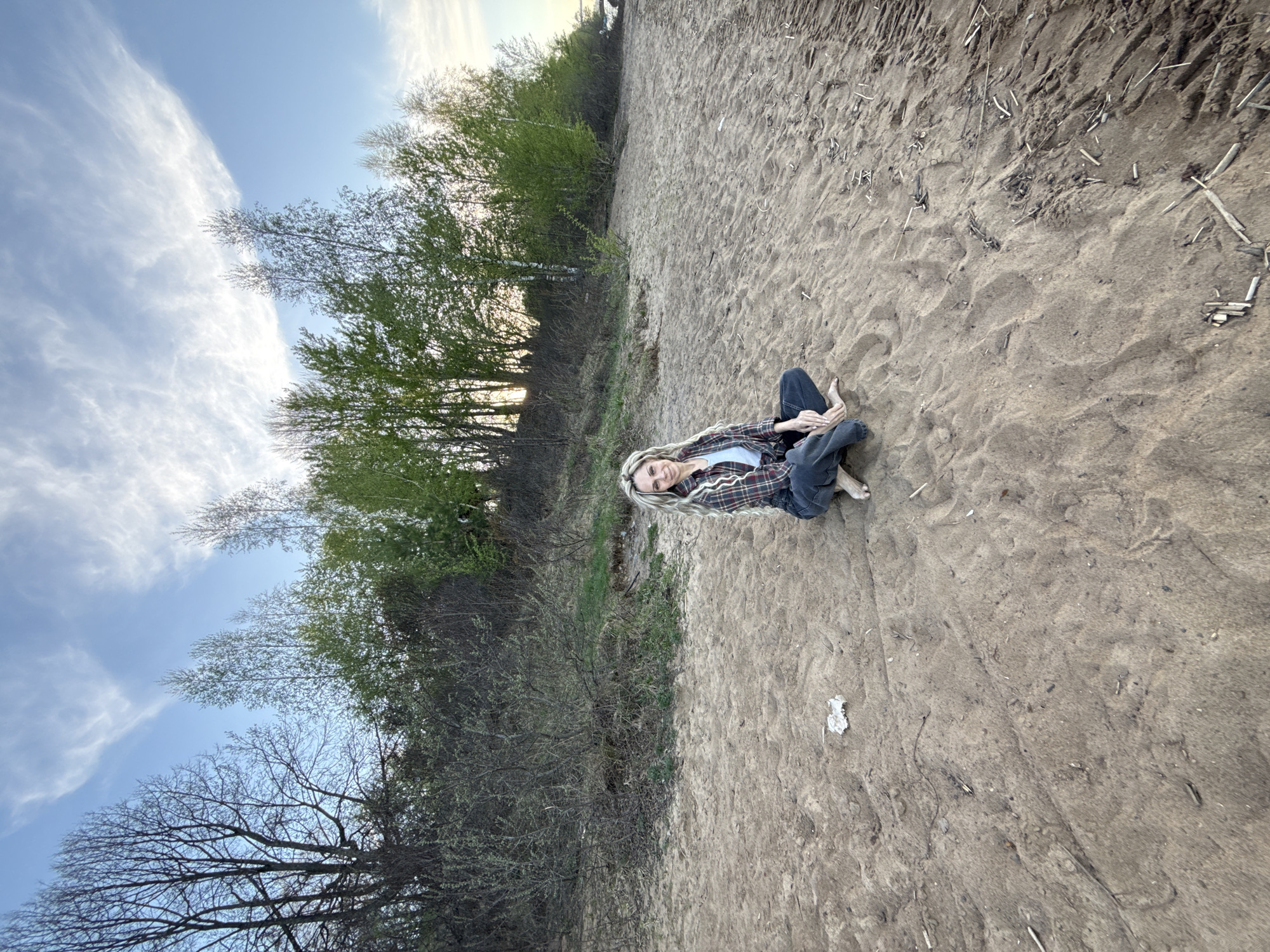 A woman with blonde hair sits on a sandy beach next to a wooded area. The photo appears to be taken outdoors in Rusanovo, Russia, as indicated by the location mentioned. She is barefoot and wearing jeans, a white top, and a plaid shirt. The background includes trees with varying degrees of foliage. A light blue sky with wispy clouds is visible. Debris is scattered on the sand.