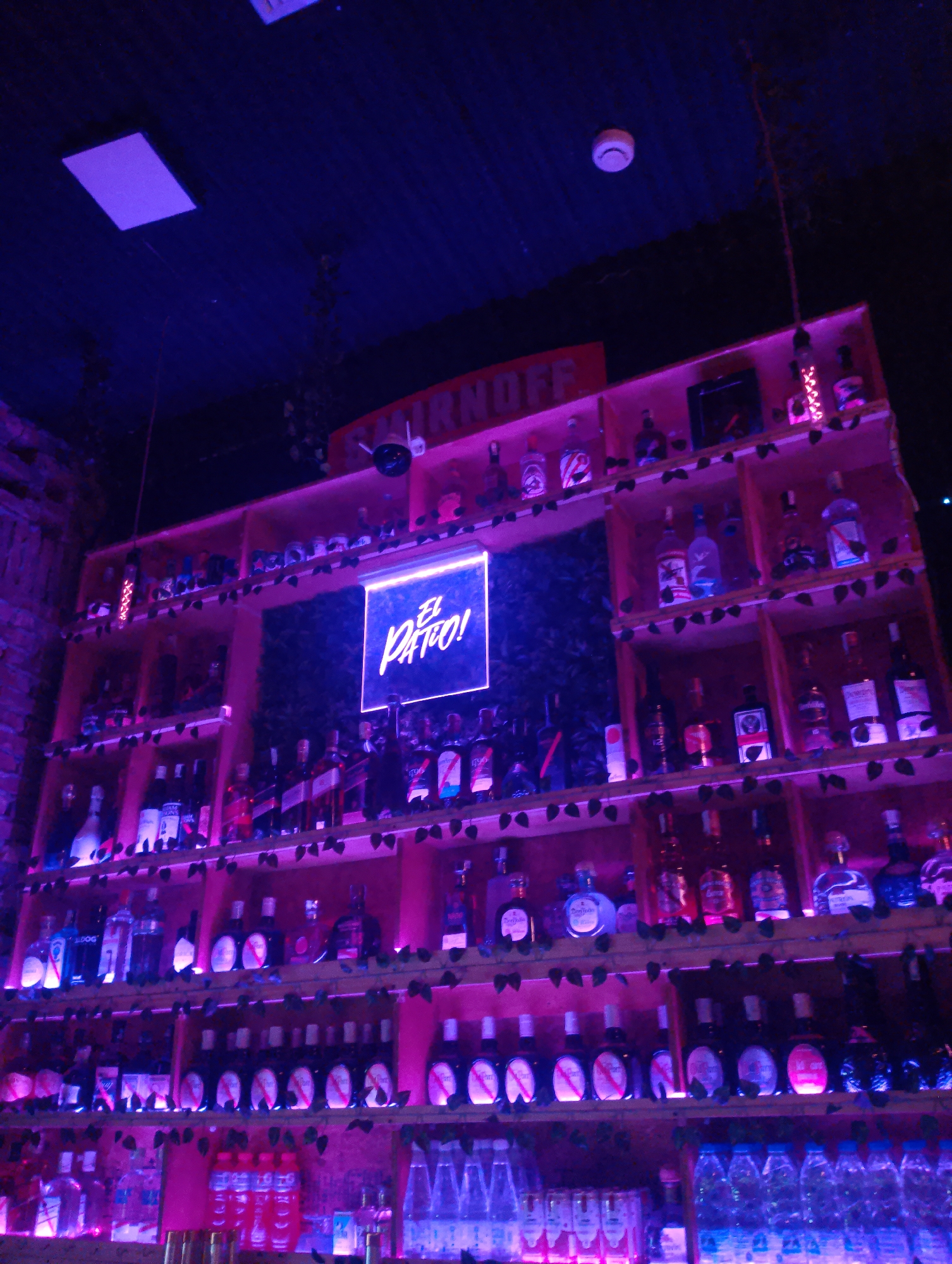 The image shows a well-stocked bar shelf under purple and blue neon lighting. The bar is called "El Patio!", as seen in a brightly lit sign. On the top of the bar is the word "SMIRNOFF". The shelves are stocked with a variety of bottles of alcohol and mixers. The shelves are decorated with faux greenery. Overhead there is a square light and a smoke detector. Water bottles are lined along the bottom shelves. The bar is located in the city of Porlamar, Venezuela. There are no people visible in the image.