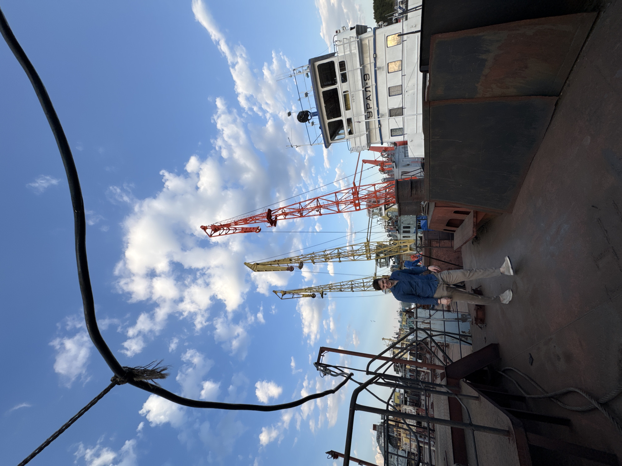 The image shows a man standing on a rusted metal surface, likely the deck of a ship or barge, with a blue sky and scattered clouds in the background. He wears a blue shirt, khaki pants, and white shoes. Several cranes are visible behind him, along with other ships and structures. Part of a large white ship with the name "УРАЛ-9" is visible to the right of the frame. A thick black cable is prominent in the foreground, partly obscuring the view. This is in the city of Chaikovsky, Russia.